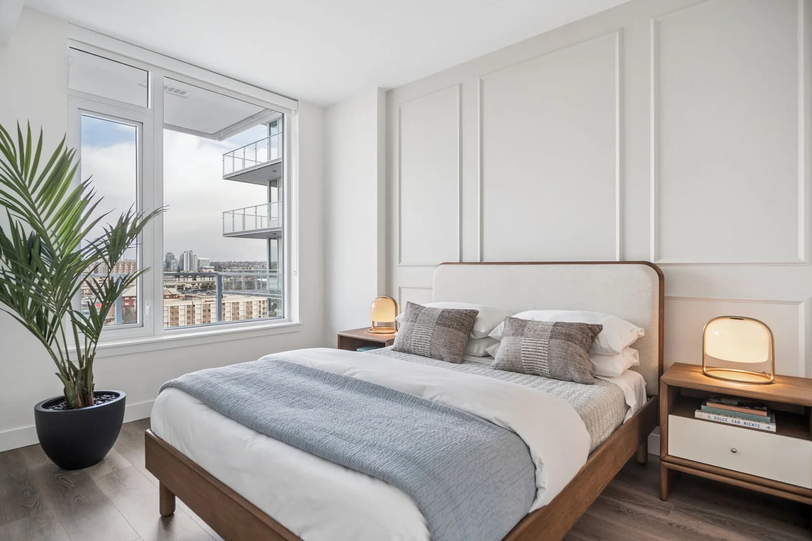 Milano at Capstan Village – A modern, bright bedroom features a wood-framed bed dressed in crisp white linens and a muted blue throw, set against a white wall with subtle picture frame molding. Large floor-to-ceiling windows next to a potted palm plant offer a view of neighbouring modern balconies and distant city buildings.