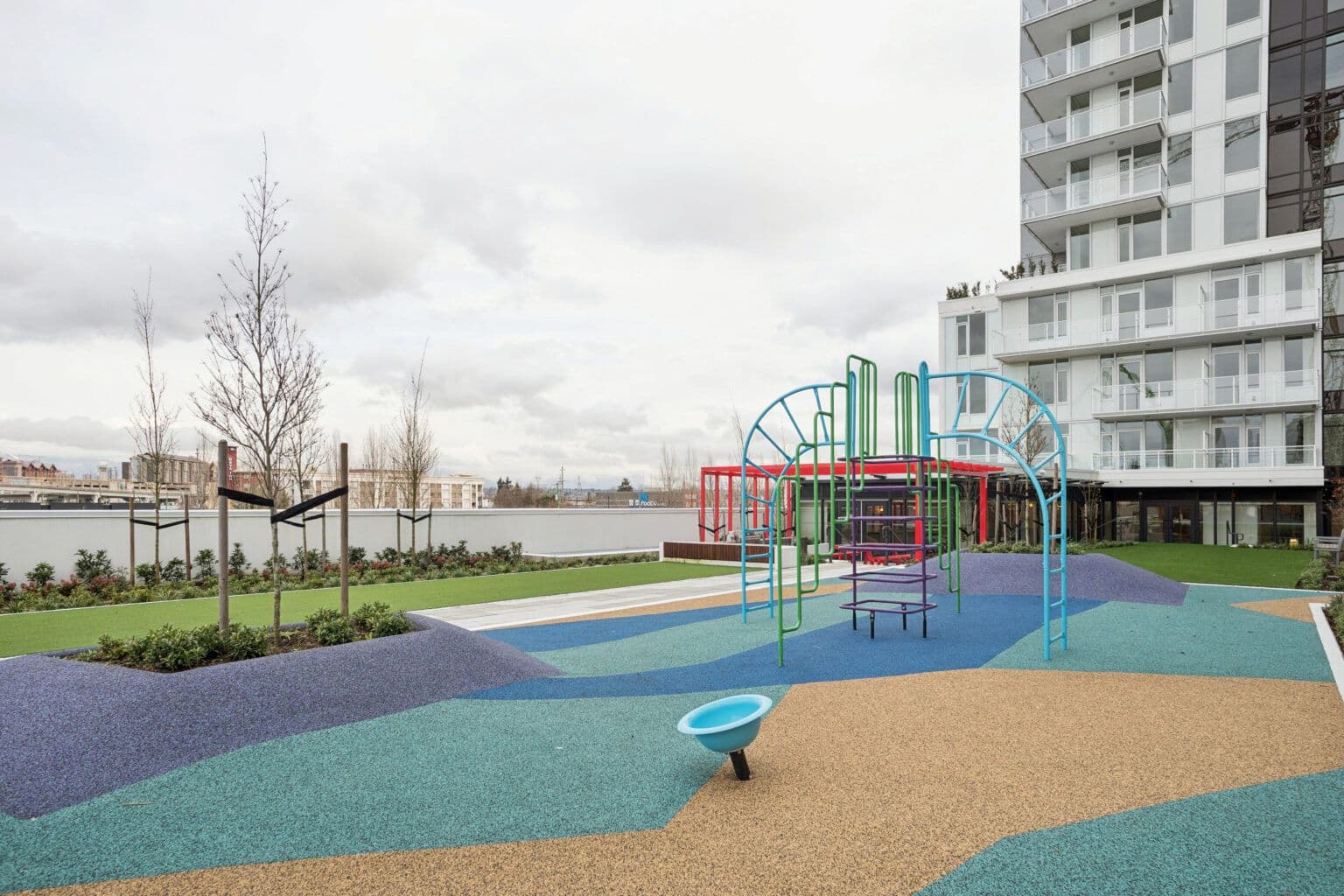 Milano at Capstan Village – An outdoor children's play area features colorful climbing structures and a vibrant, cushioned safety surface set on a podium deck. Next to the playground are manicured lawn patches, young trees, and the base of a modern residential high-rise tower under an overcast sky.