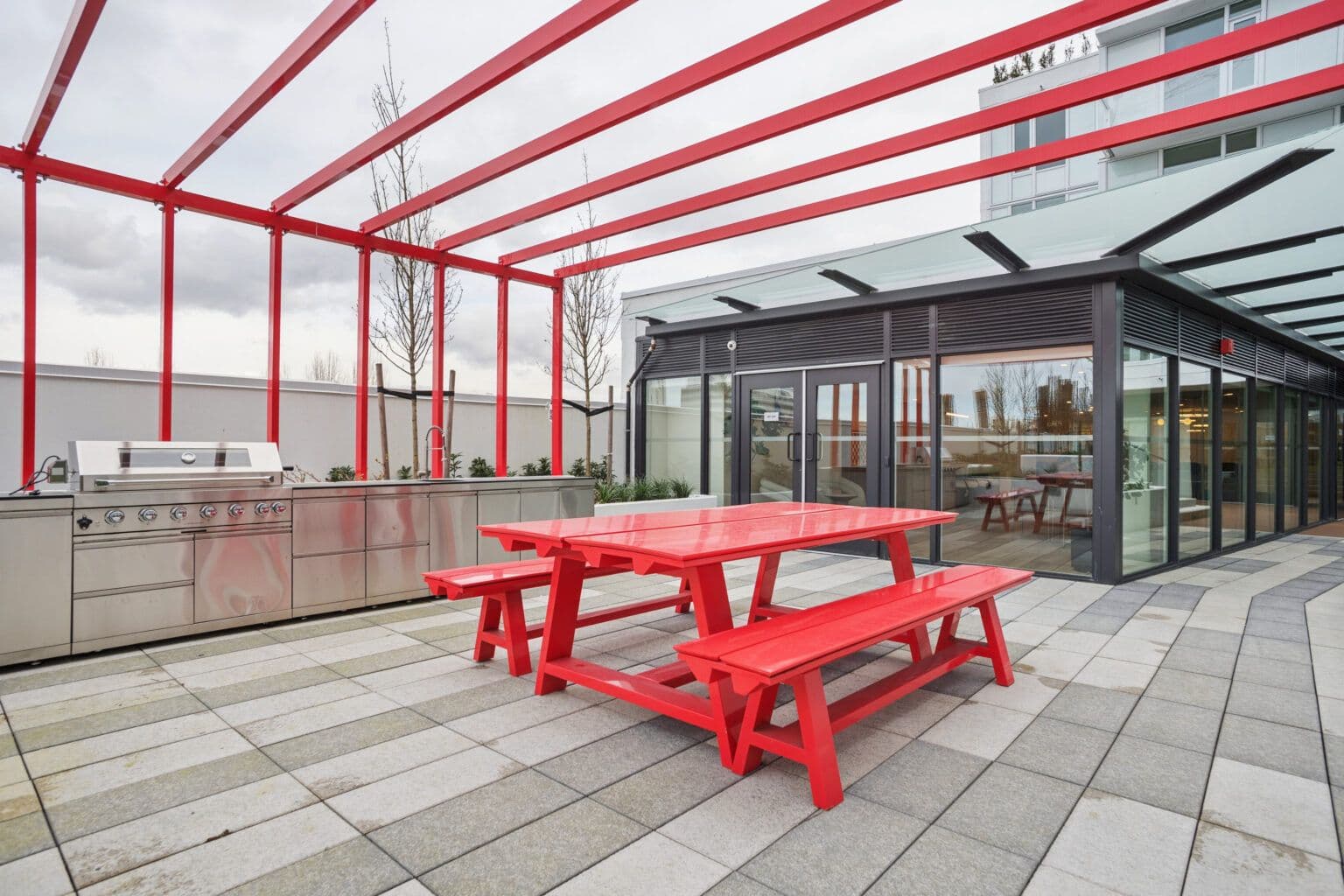 Milano at Capstan Village – An outdoor rooftop terrace features a stainless steel BBQ cooking station and a bright red picnic table positioned beneath an open-frame red pergola. The paved patio is situated adjacent to a modern glass-enclosed indoor lounge under an overcast sky.