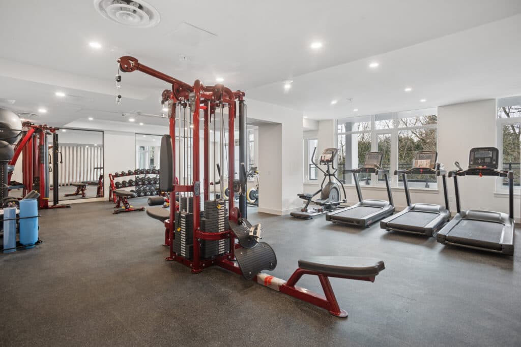 Milano at Capstan Village – A bright residential fitness facility featuring red multi-station cable machines, a rack of dumbbells, and a line of treadmills facing large windows. The space has dark rubber flooring, white walls, recessed ceiling lights, and mirrors reflecting the gym layout.