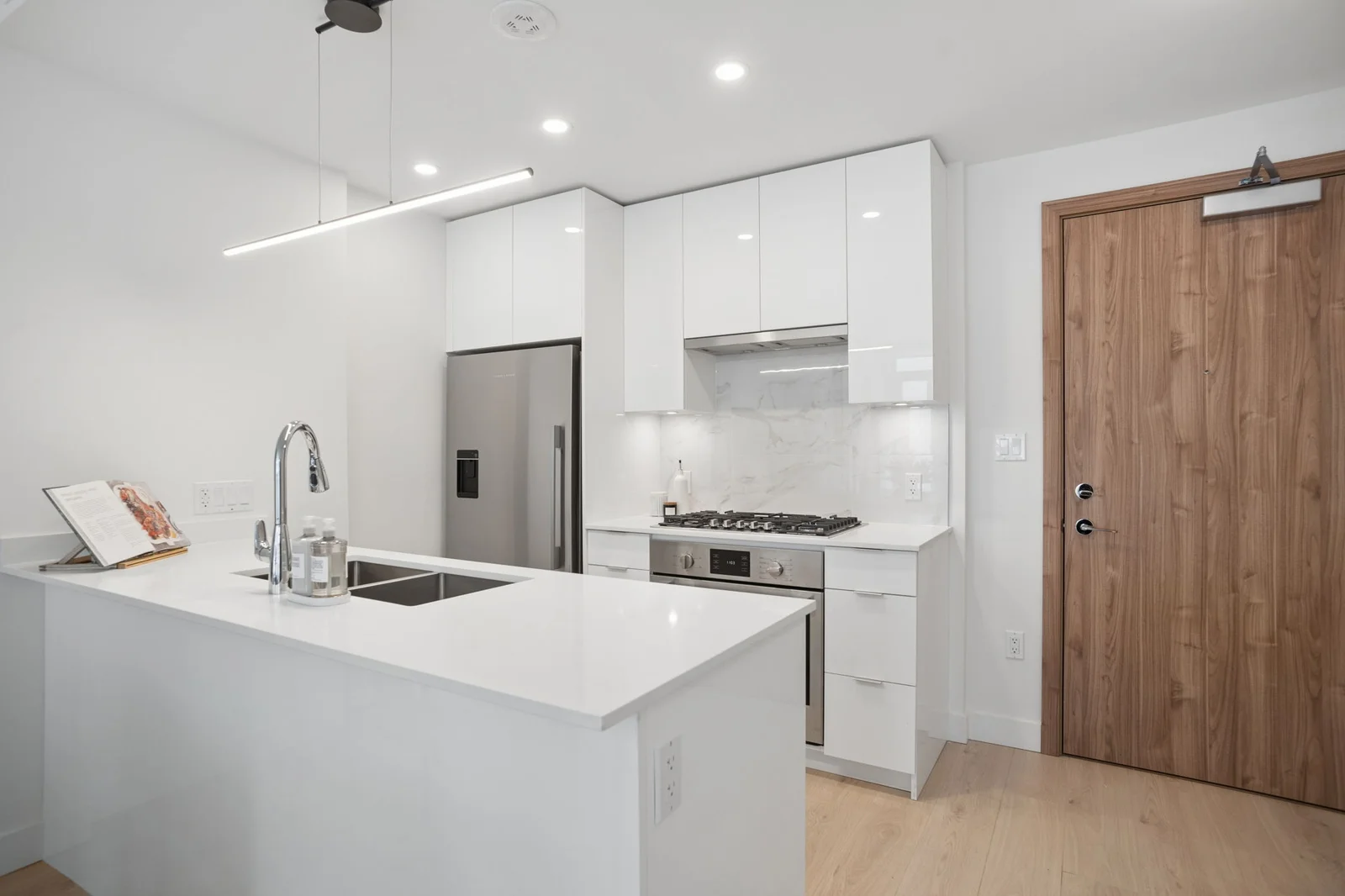 Milano at Capstan Village – A modern, bright condo kitchen featuring glossy white cabinetry, white quartz countertops, and stainless steel appliances. A kitchen island with an undermount double sink sits under a minimalist linear LED pendant light, with a warm wood entryway door visible to the right.