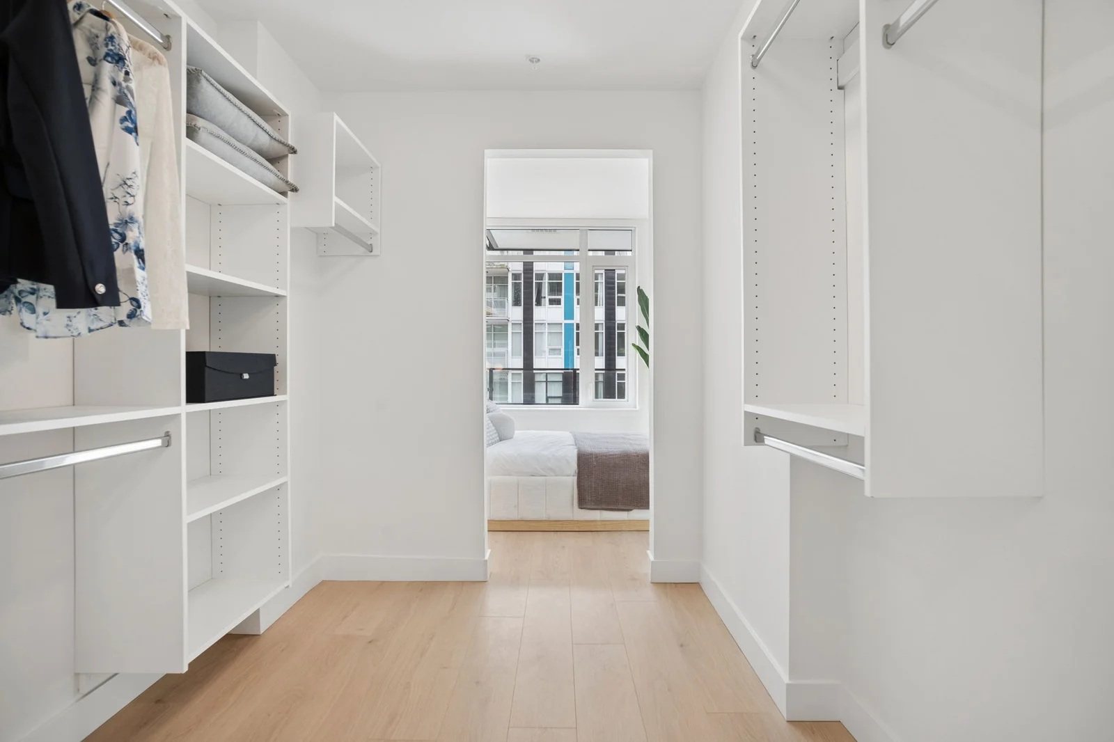 Milano at Capstan Village – A bright, custom walk-in closet featuring all-white built-in shelving, hanging rods, and light wood flooring. An open doorway at the center provides a view straight into an adjacent bedroom flooded with natural light from large windows.