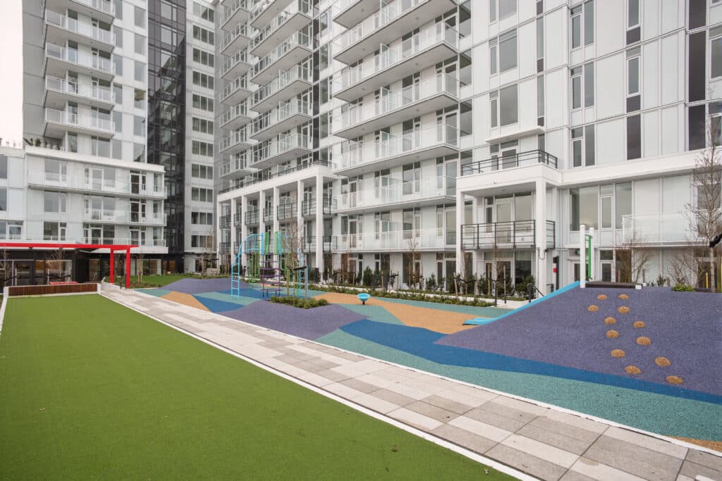 Milano at Capstan Village – A modern outdoor courtyard featuring a colorful, rubberized play area with climbing structures and slides, a paved walkway, and a green lawn area. The space is enclosed by a tall, white modern residential condominium tower with numerous glass balconies.