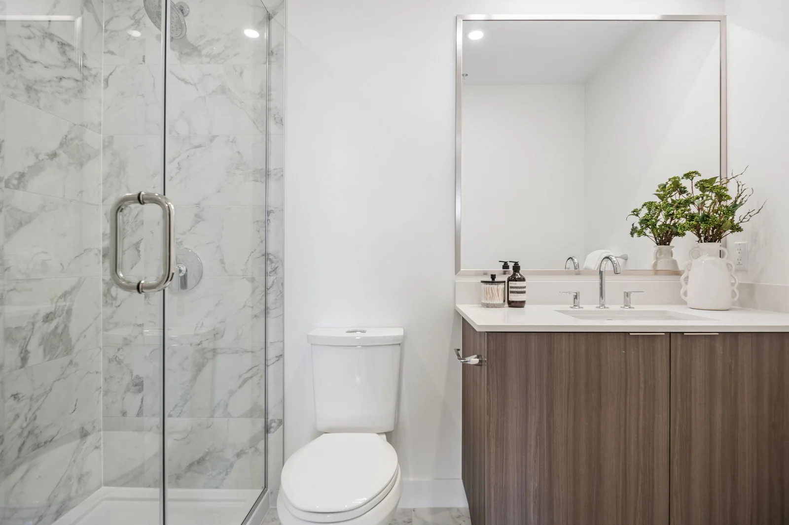 Milano at Capstan Village – A bright, modern bathroom featuring a walk-in glass shower lined with gray-and-white marble-patterned tiles. A dark wood vanity with a white countertop, a large framed mirror, and a white toilet complete the clean, contemporary space.