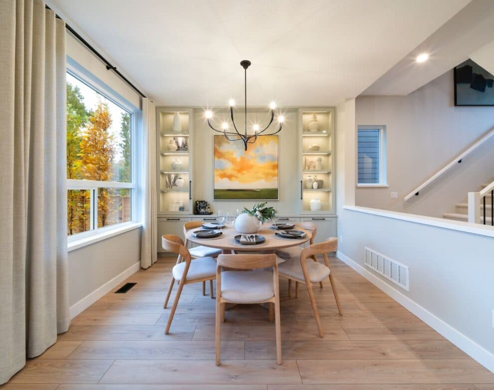 South Hill – Dining room with a round wood table and upholstered chairs, a black sculptural chandelier, built-in lit display shelving and a large window with autumn trees outside.
