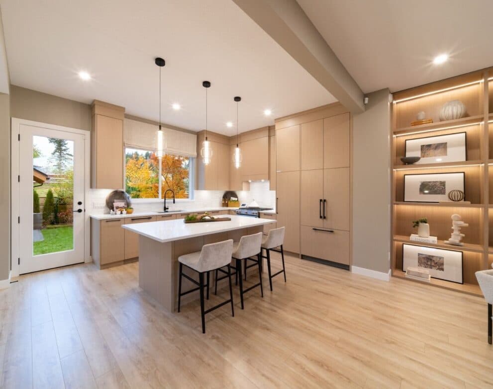 South Hill – Open-concept kitchen with light wood cabinetry, a large island with seating, pendant lighting, built-in lit display shelves and a door to the backyard.