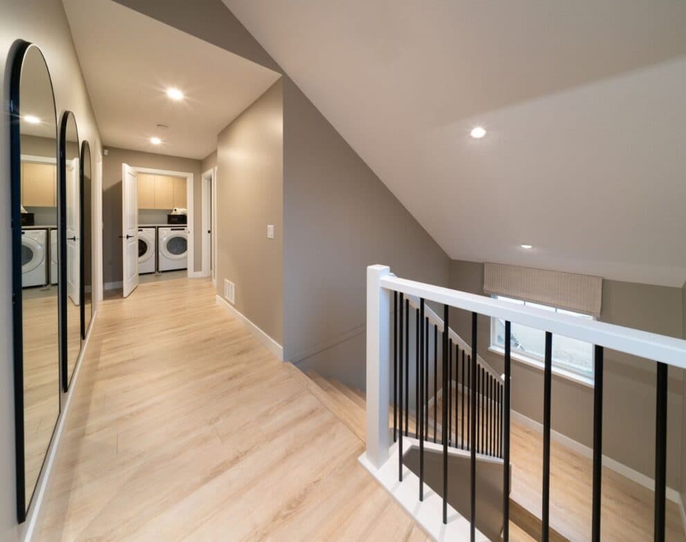 South Hill – Upper hallway with a full-length mirror, wood flooring, a black-railed staircase, and an open laundry room with stacked/side-by-side washer and dryer.