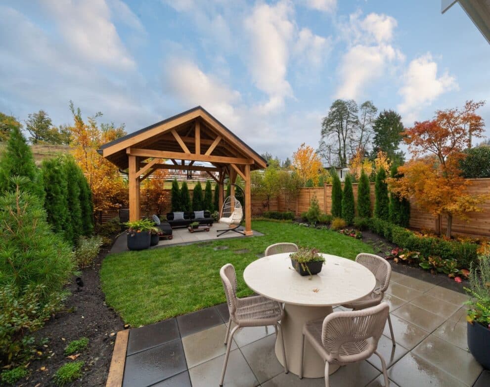 South Hill – Backyard patio with a round outdoor dining table and woven chairs in the foreground and a timber-framed gazebo with lounge seating and a hanging egg chair beyond, surrounded by autumn-colored landscaping.