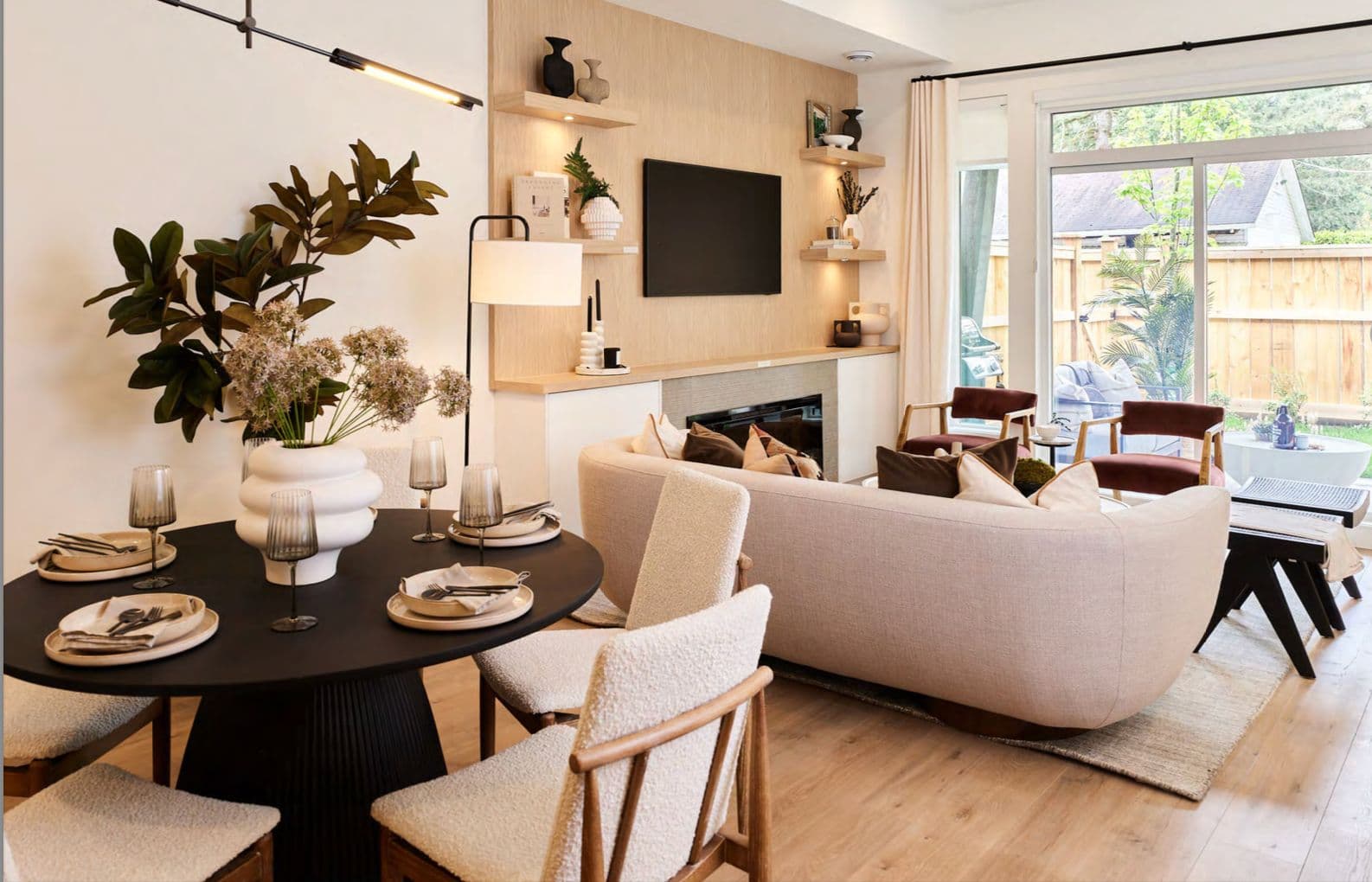 Everwood – Living and dining area with a round black dining table, cream boucle dining chairs, a beige sofa, fireplace with wall-mounted TV, and sliding doors to a backyard patio.