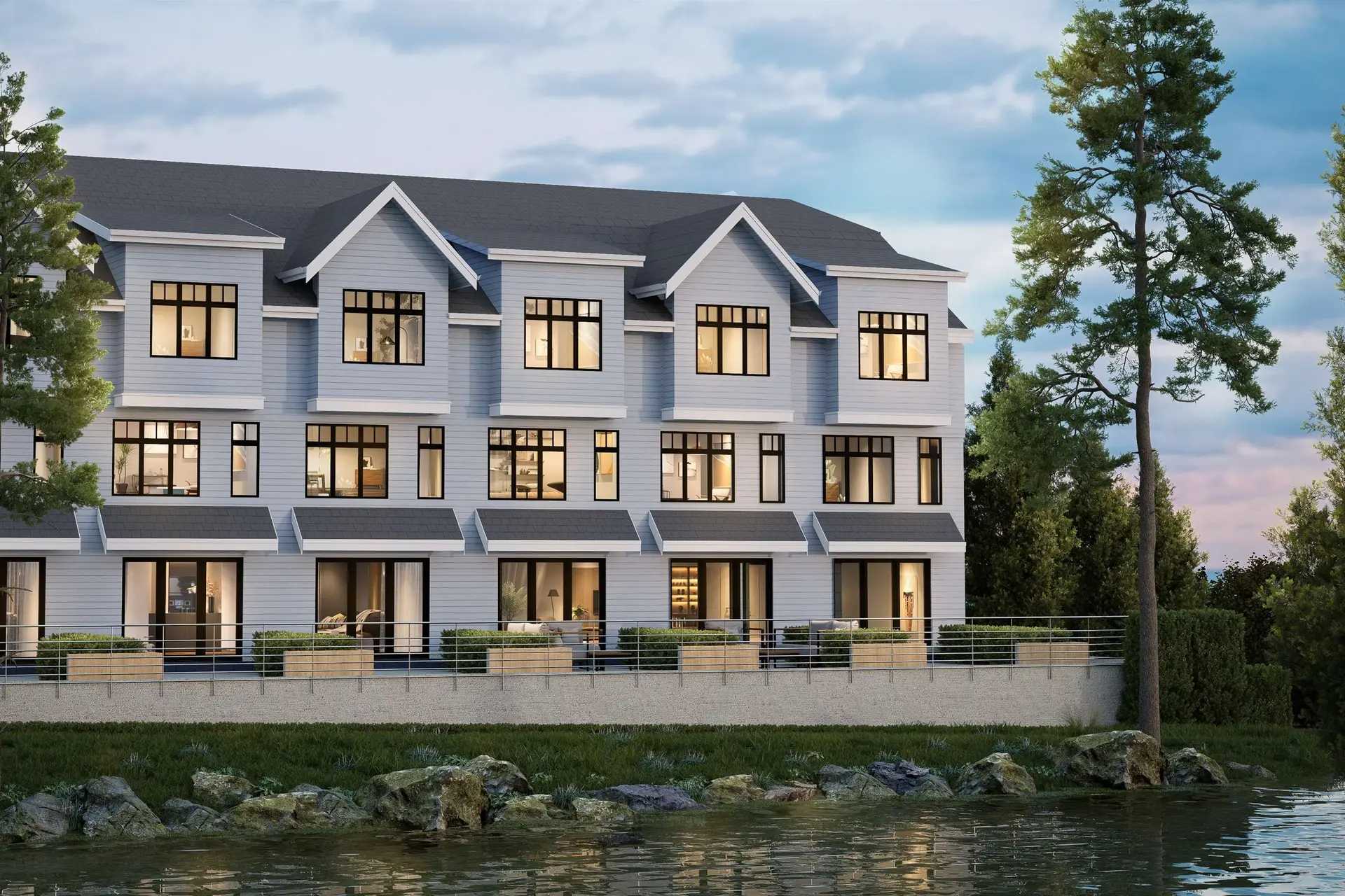 Holland Row II – Evening waterfront view of townhomes with lit interiors, patios along a rocky shoreline, and a large pine tree in the foreground.