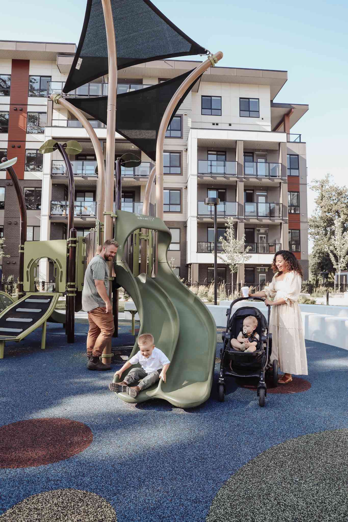 York Residences - Building C – A father and mother with two young children playing on a green slide at an outdoor playground, with the condo building visible behind them.