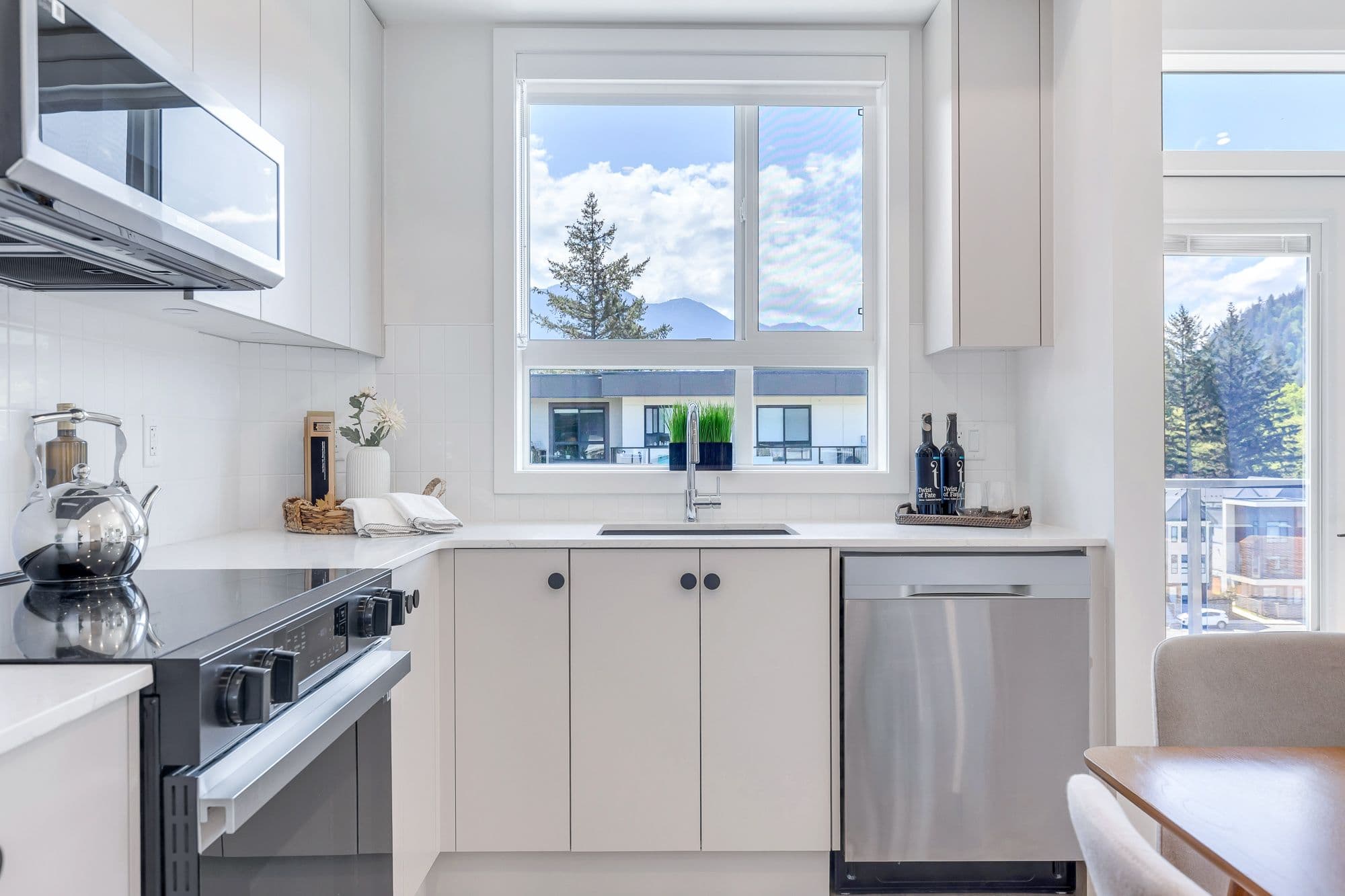 York Residences - Building C – A white kitchen with an L-shaped counter, stainless dishwasher, and a large window above the sink showing mountain and neighbouring rooftop views.