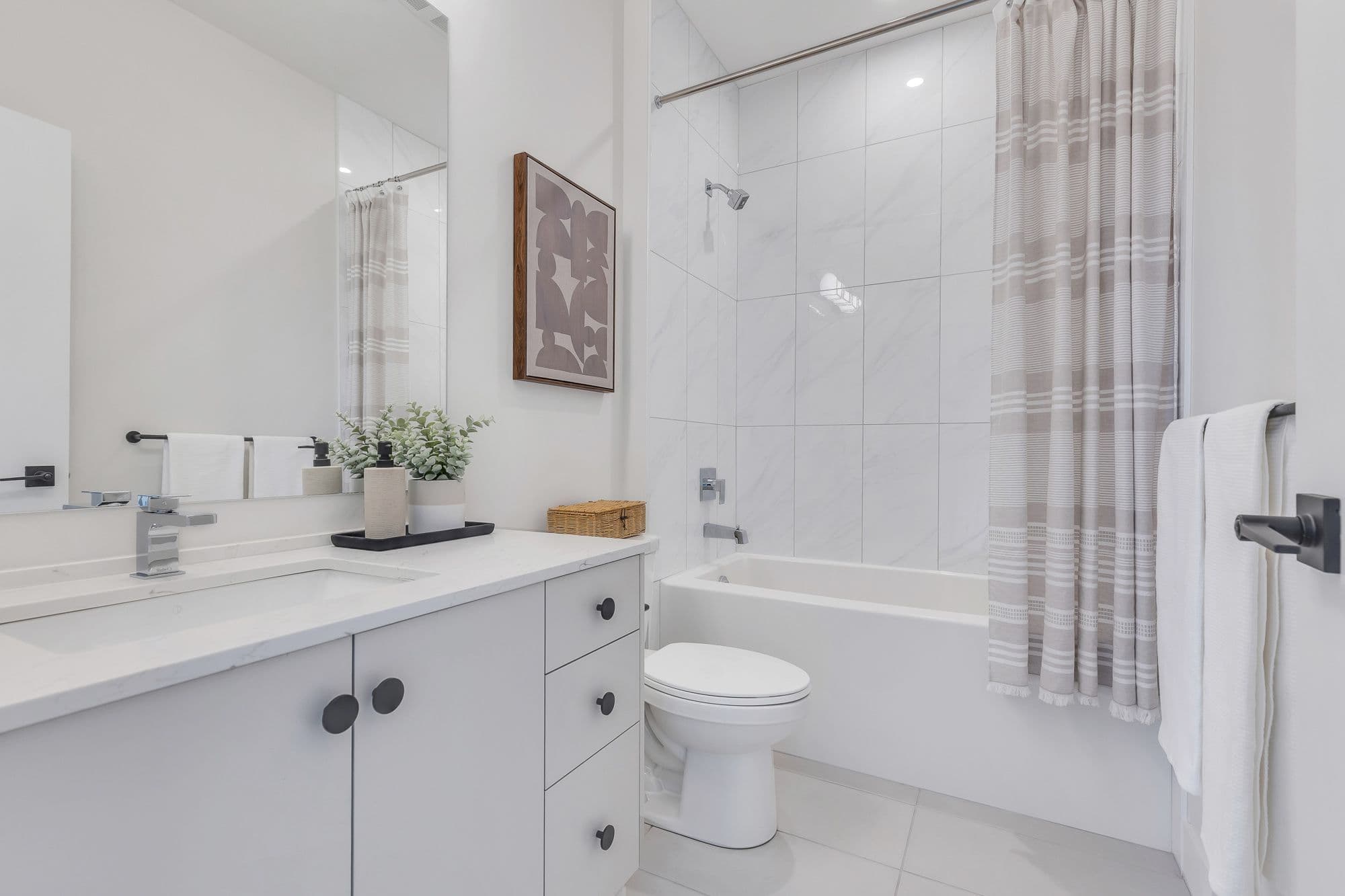 York Residences - Building C – A bathroom with a double vanity, framed abstract art, and a tiled tub/shower combo with a striped shower curtain.