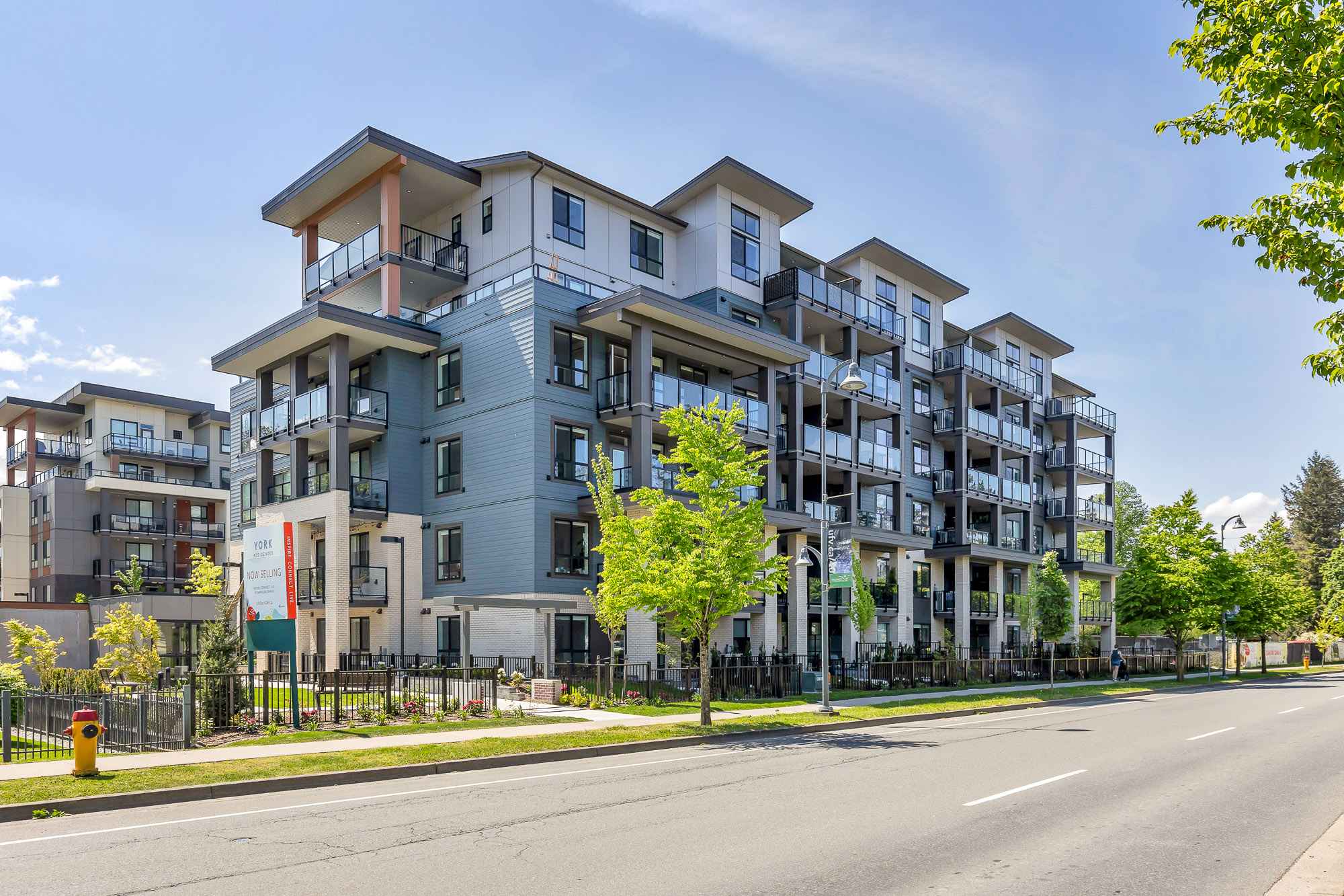 York Residences - Building C – Street view of a mid-rise condo building with blue-grey and white siding, multiple balconies, and a 'York Residences - Now Selling' sign out front.