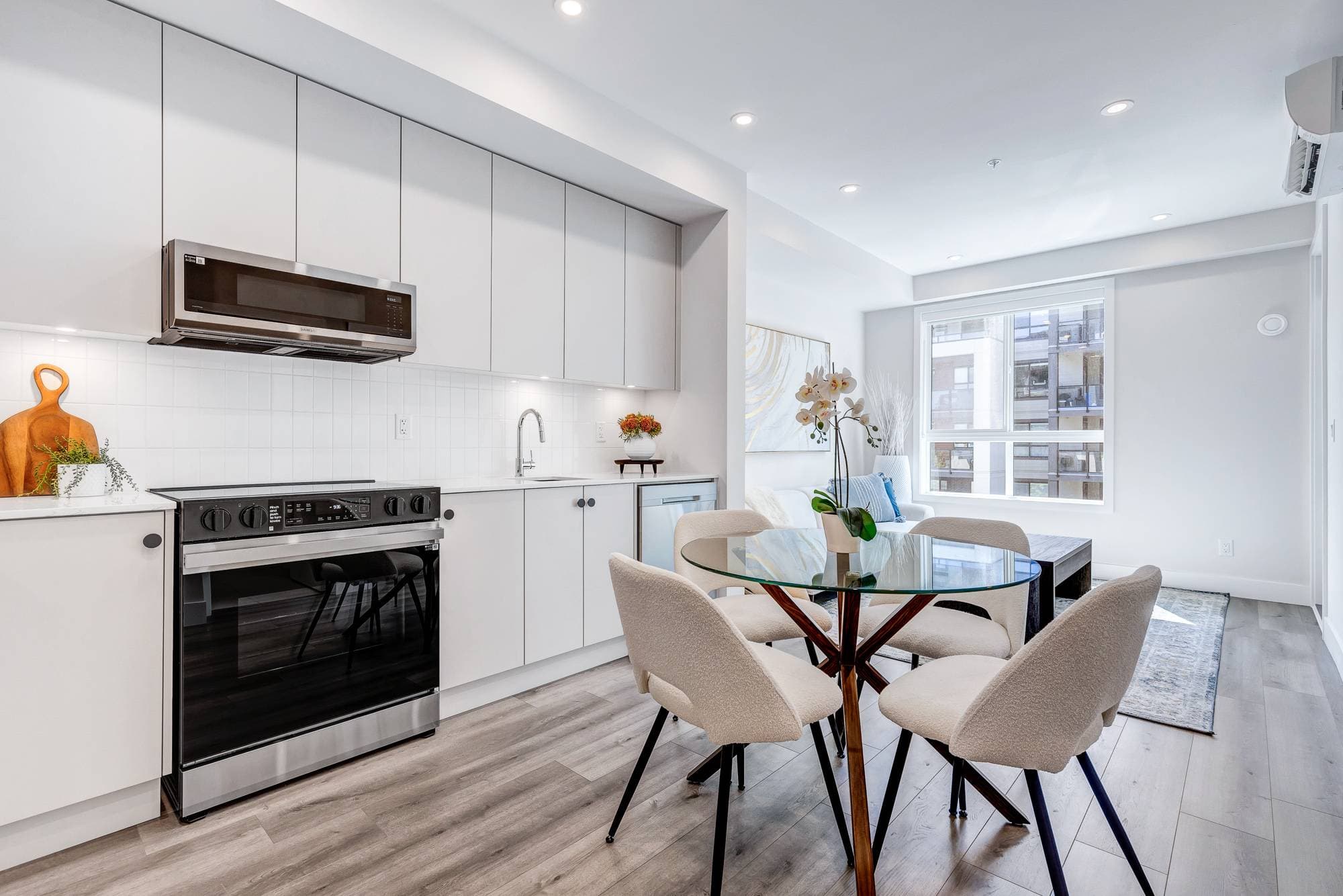 York Residences - Building C – An open-concept kitchen and dining area with white cabinetry, a round glass dining table, cream chairs, and a living room visible beyond.