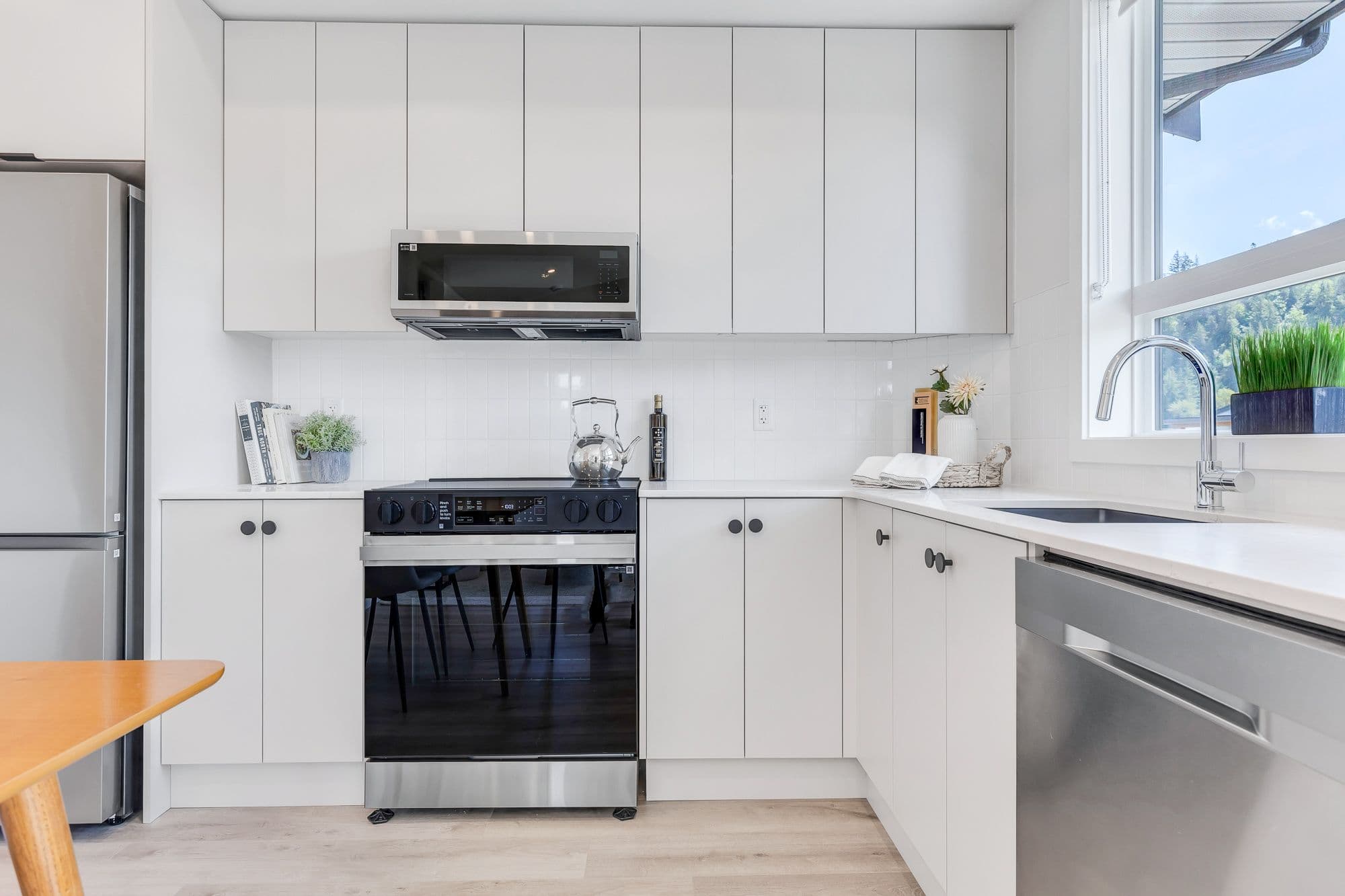 York Residences - Building C – A white kitchen with tall cabinetry, a stainless range and microwave, and a window looking out to trees and hills.