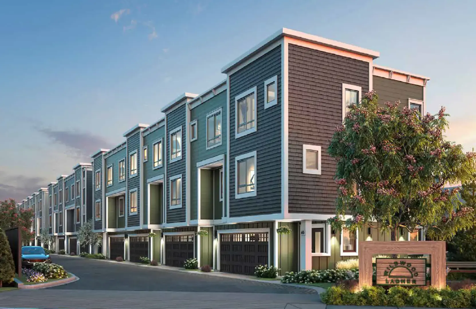 Wildwood Ladner – A row of modern three-story townhomes clad in dark grey, teal, and brown shake siding stands along a paved internal drive under a bright dusk sky. Ground-floor garages, crisp white trim, manicured landscaping, and an illuminated community signage monument complete the inviting residential streetscape.