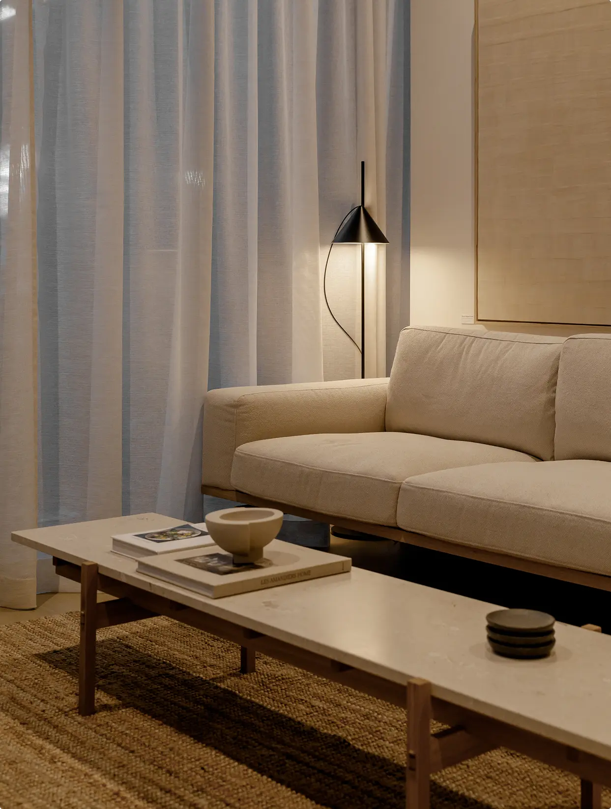 Söenhaus – Showroom-style living room corner with a beige linen sofa, sheer curtains, a black cone floor lamp, and a stone coffee table with books on a jute rug.