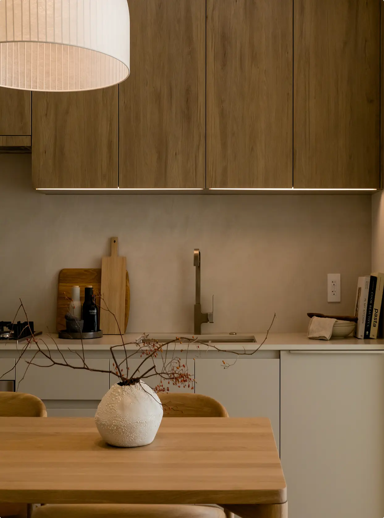 Söenhaus – Close-up of a kitchen counter and backsplash with wood upper cabinets, a wall-mount faucet, cutting boards, and a dining table with a vase of branches in the foreground.