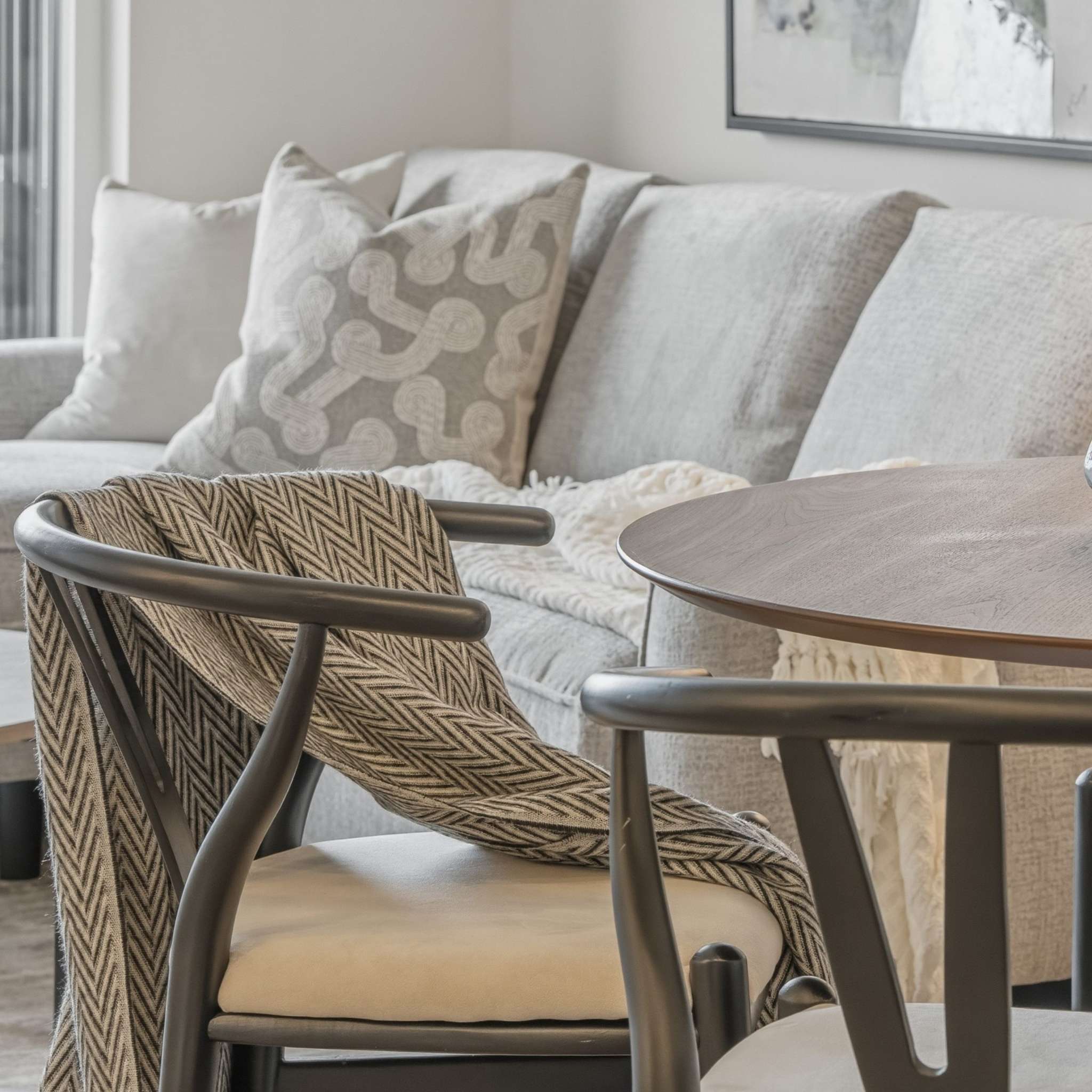 City Corners – A cozy, close-up view of a living and dining area featuring a modern dark wood dining chair draped with a chevron-patterned throw blanket beside a round wooden table. In the background sits a comfortable light gray sofa dressed with patterned cushions and a soft knit blanket, illuminated by soft natural daylight.