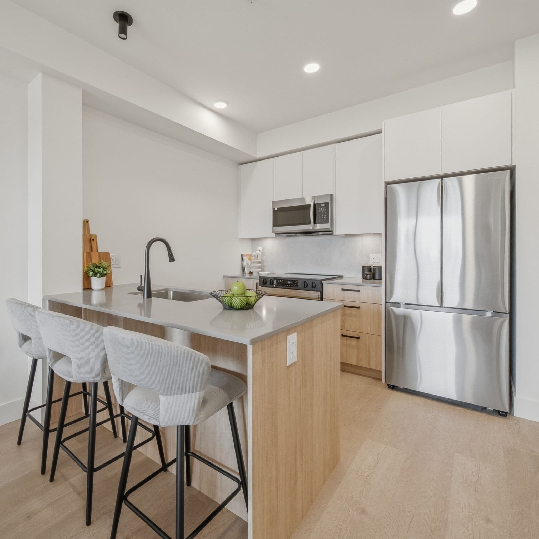 City Corners – A modern open-concept kitchen features a light wood island with grey quartz countertops, three fabric-upholstered barstools, and an undermount sink with a matte black faucet. The space is fitted with two-tone white and wood cabinetry, full-size stainless steel appliances including a French door refrigerator, and light wood flooring illuminated by recessed ceiling lights.