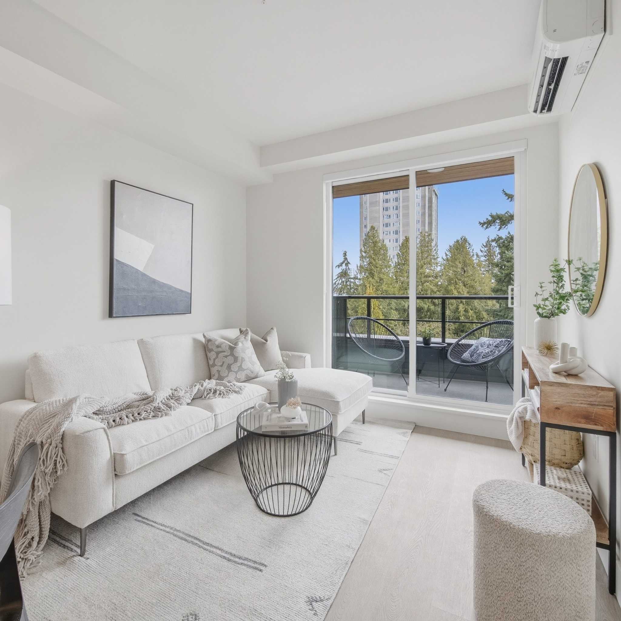 City Corners – A bright, neutrally styled living room features a white sectional sofa, a black wire-frame coffee table, and light wood flooring. Large sliding glass doors lead to a private balcony with outdoor seating that overlooks lush green treetops and a high-rise building under a clear blue sky.
