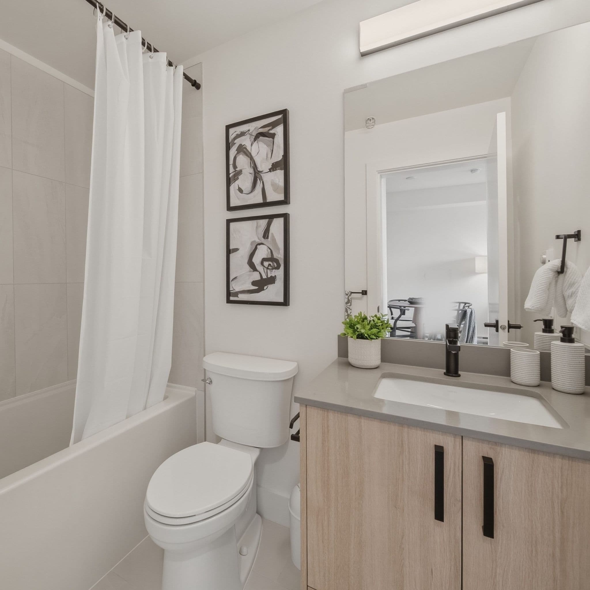 City Corners – A clean, modern bathroom featuring a light wood vanity cabinet with gray countertops and matte black fixtures alongside a standard white toilet and bathtub. The walls are finished in neutral off-white with two framed abstract art prints, a large vanity mirror, and soft interior lighting.