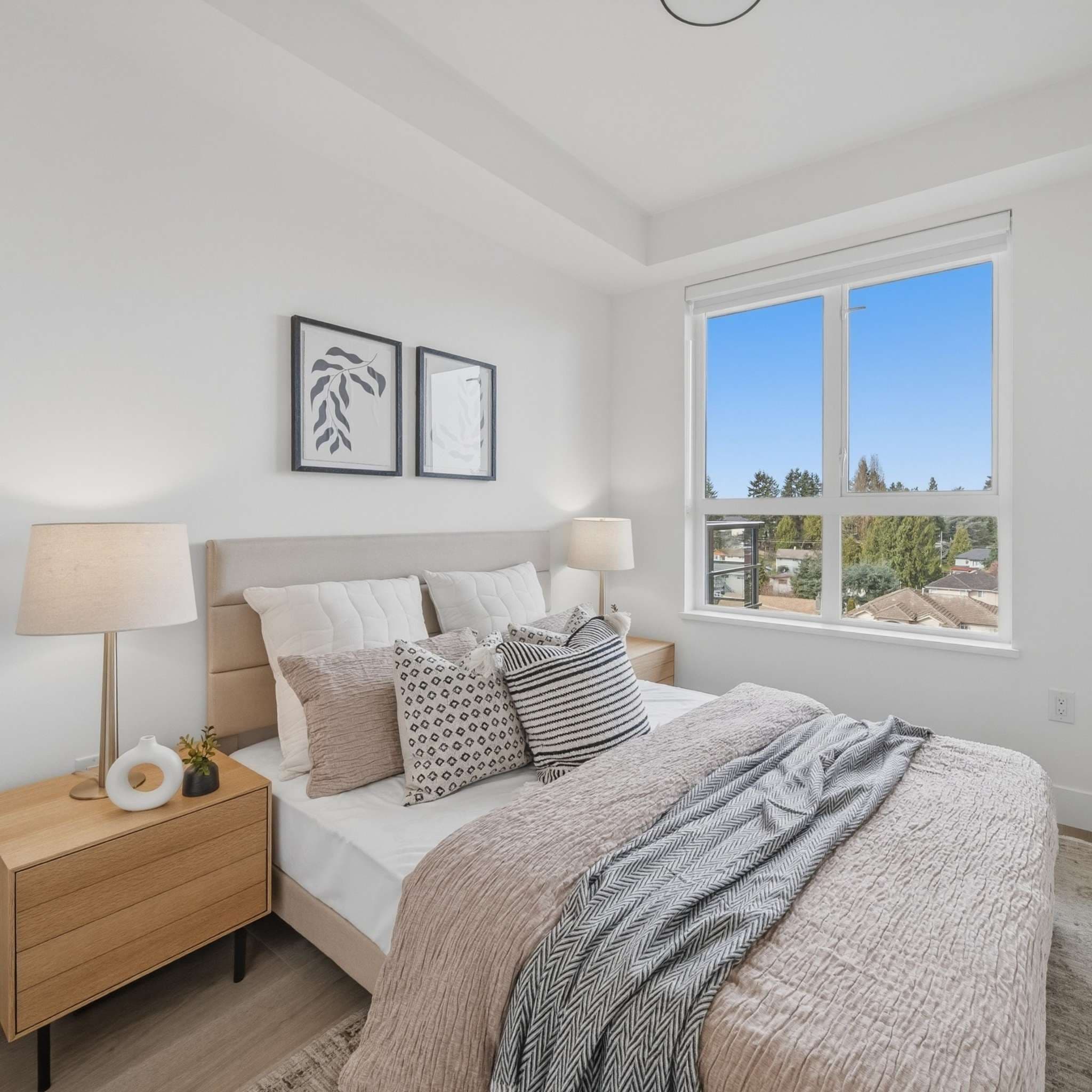 City Corners – A bright, modern bedroom featuring a neatly made bed with layered pillows, light wood nightstands with lamps, and framed minimalist botanical art on the white wall. A large window overlooks green treetops and neighbouring residential rooftops under a clear blue daytime sky.
