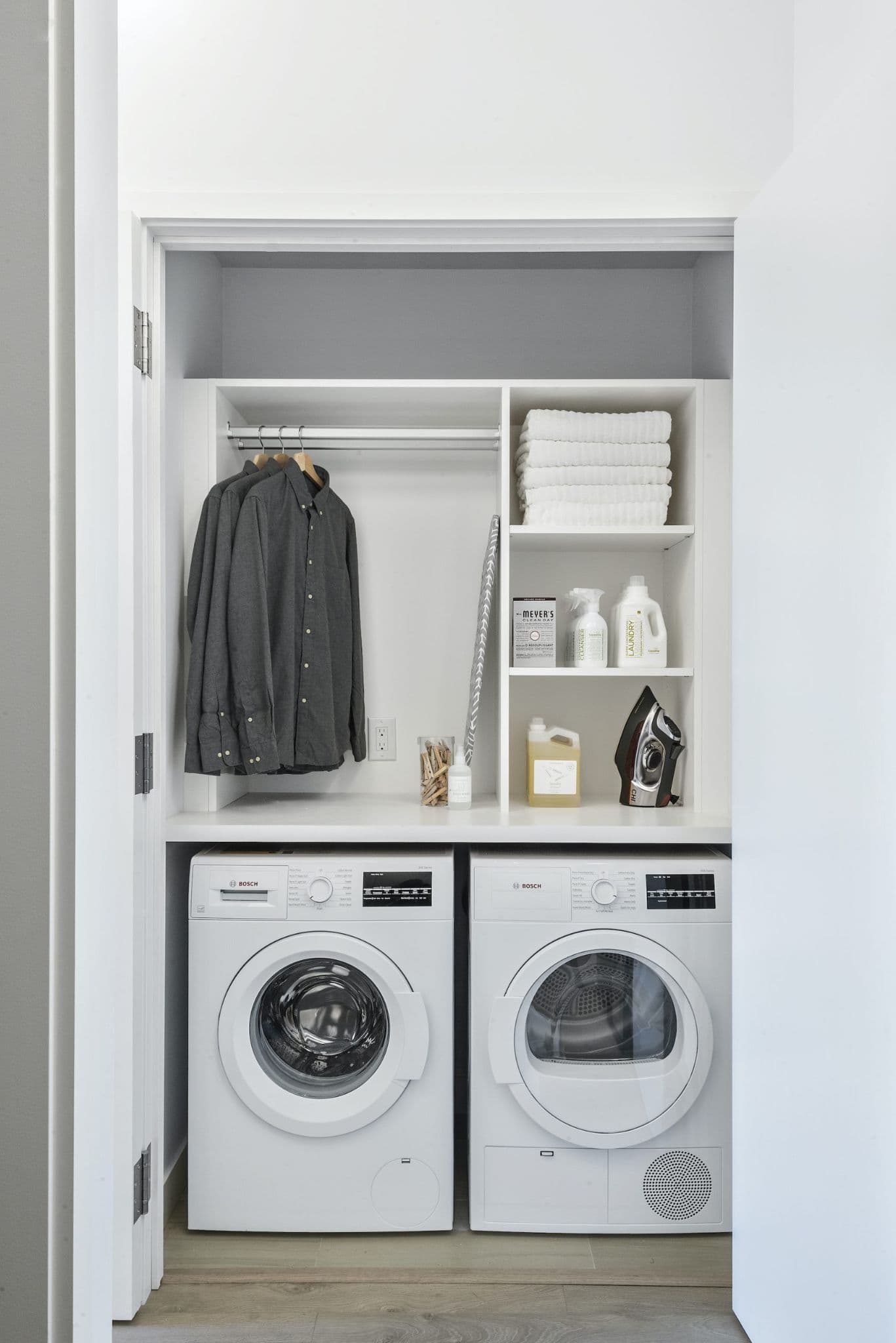 RC at CF Richmond Centre – A clean, modern laundry closet features side-by-side white Bosch front-loading washer and dryer units beneath custom white shelving. The upper storage area includes a small hanging rod with collared shirts, folded white towels, laundry supplies, an iron, and a folded ironing board.