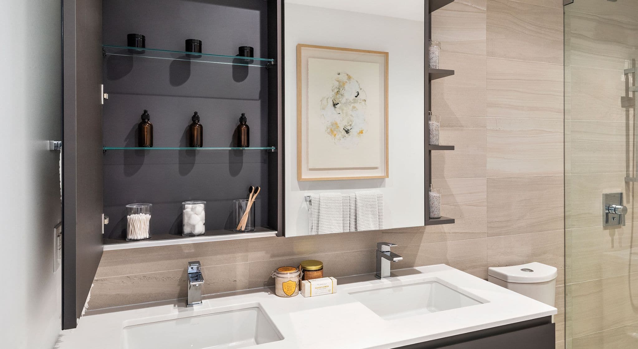 RC at CF Richmond Centre – A modern bathroom vanity features dual undermount sinks set in a white countertop, chrome fixtures, and a large mirror flanked by framed abstract artwork and open storage shelves holding toiletries. Beige travertine-style wall tiles extend past a glass shower enclosure on the right, creating a bright, spa-like atmosphere.