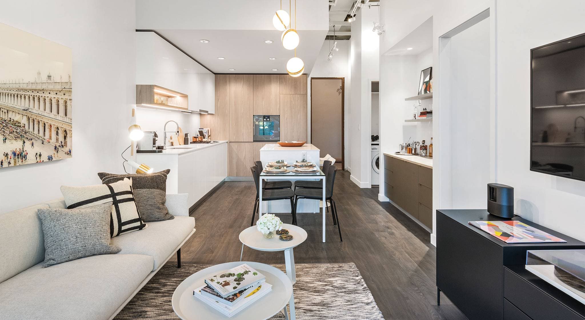 RC at CF Richmond Centre – A modern open-concept condo interior featuring a bright living room with a light grey sofa that leads into a sleek kitchen with minimalist white and light wood cabinetry. The space includes a small dining table set for four near the kitchen island, dark hardwood floors, high ceilings with track lighting, and a contemporary wall-mounted television.