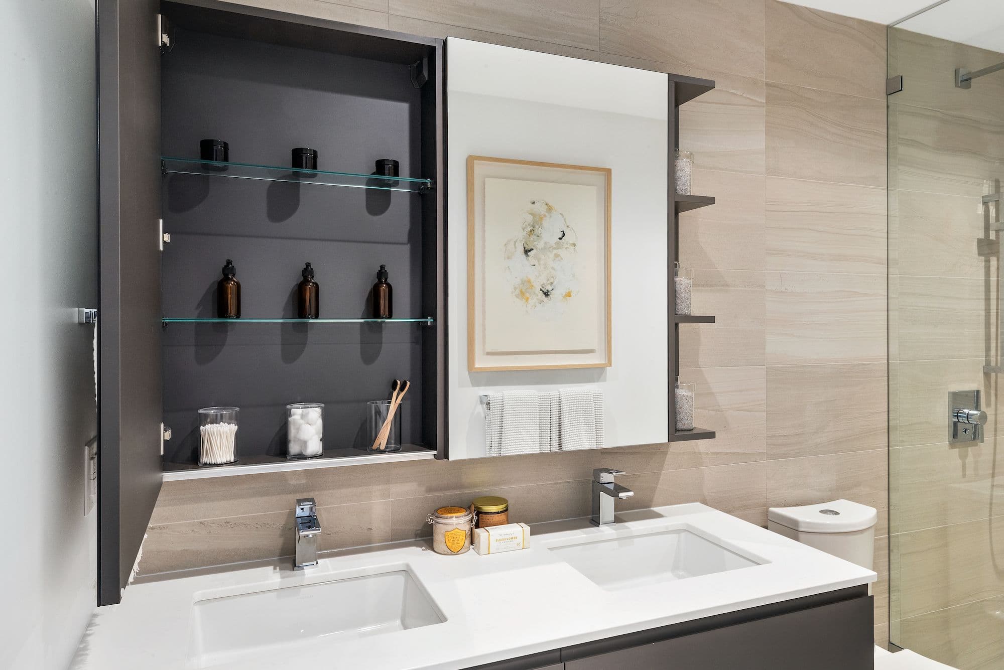 RC at CF Richmond Centre – A modern bathroom vanity features dual undermount sinks set into a white quartz countertop, framed by beige large-format tile walls. An open dark gray medicine cabinet displays neatly organized glass jars and toiletries alongside a mirrored cabinet section, with a frameless glass shower enclosure visible on the right.