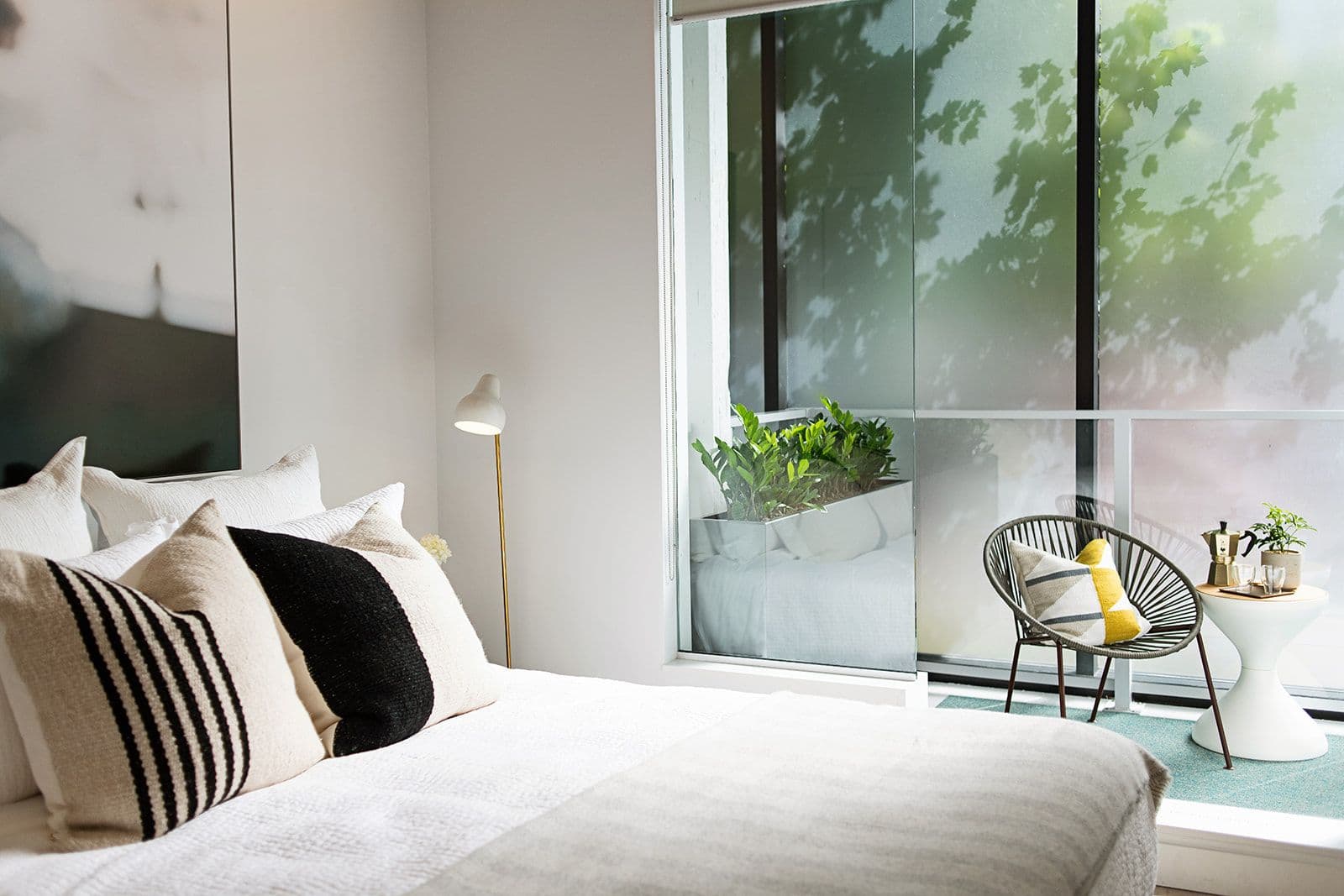 RC at CF Richmond Centre – A modern bedroom styled with textured throw pillows on a crisp white bed next to floor-to-ceiling glass doors. The doors open directly onto a private balcony featuring a woven lounge chair, small side table, potted plants, and frosted glass panels for privacy.