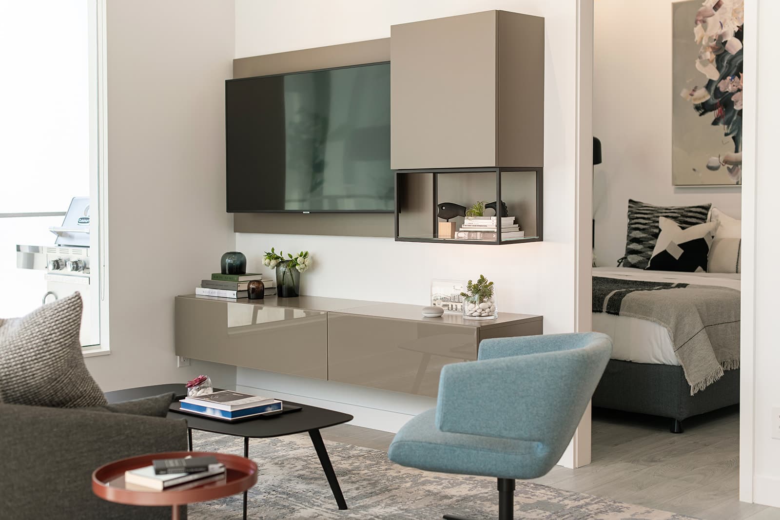 RC at CF Richmond Centre – A modern living room features a wall-mounted flat-screen television integrated into grey floating cabinetry, complemented by a light blue accent chair and dark coffee tables. An open doorway adjacent to the entertainment unit offers a view into a neatly styled bedroom with a patterned throw and abstract artwork.