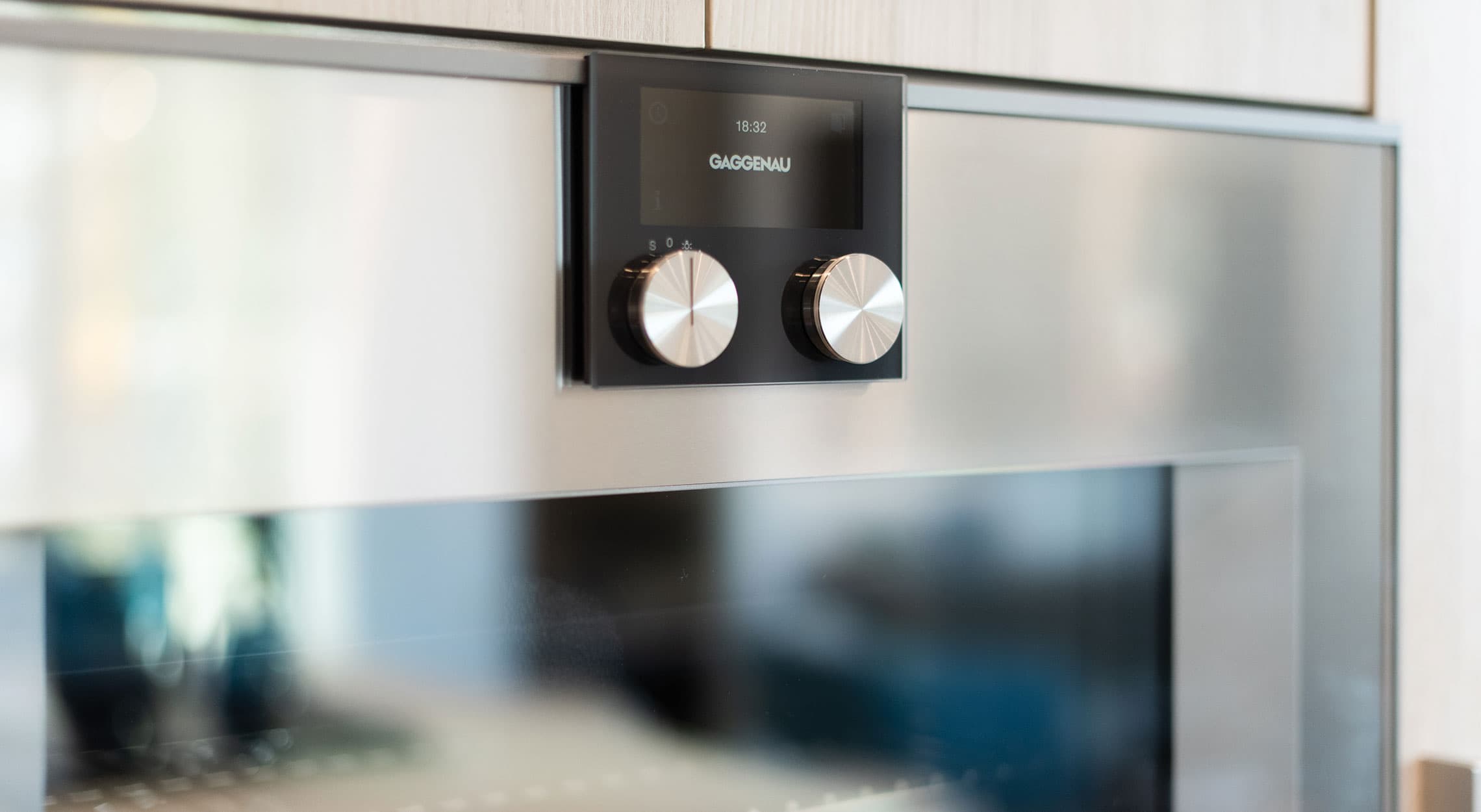RC at CF Richmond Centre – A extreme close-up shot highlights a high-end built-in Gaggenau kitchen appliance featuring sleek stainless steel surfaces and a black digital control panel with two metallic knobs. Light wood cabinetry is subtly visible along the top edge, emphasizing modern luxury finishes and clean design.
