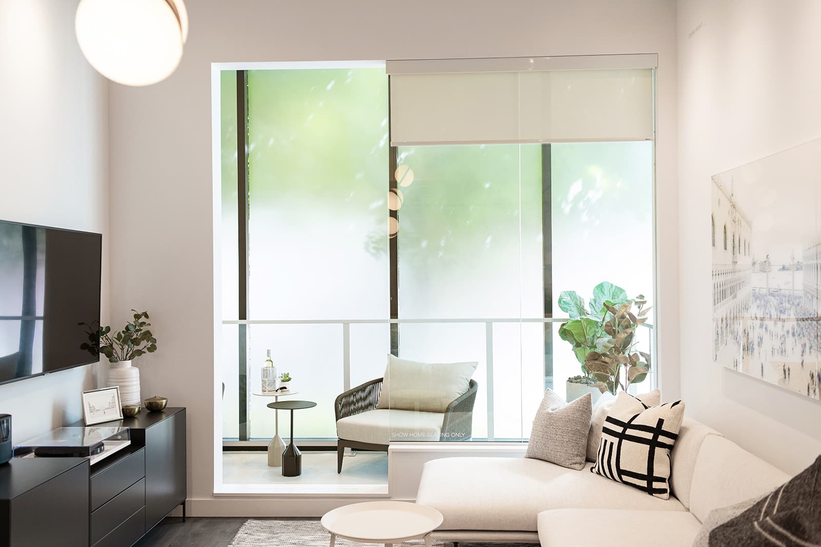 RC at CF Richmond Centre – A modern living room features a white sectional sofa, dark media console, and wall-mounted television leading out to a glass-enclosed balcony with a lounge chair and side tables. Abundant natural light fills the bright, neutral-toned space, complemented by potted greenery and frosted glass privacy panels on the outdoor deck.