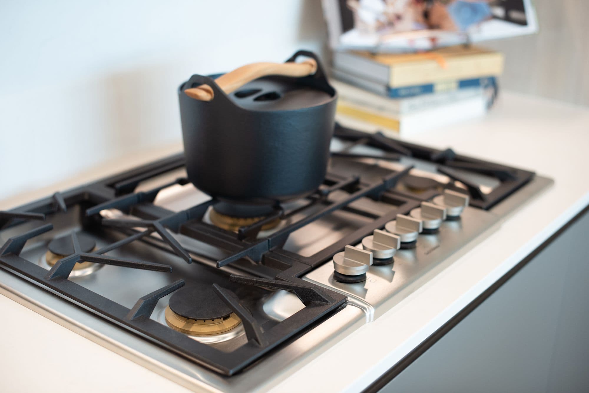 RC at CF Richmond Centre – A close-up shot features a modern stainless steel gas cooktop with heavy-duty black iron grates set into a clean white quartz countertop. A stylish black cast-iron Dutch oven with a light wood handle sits on one of the burners, with a stack of cookbooks subtly blurred in the background.