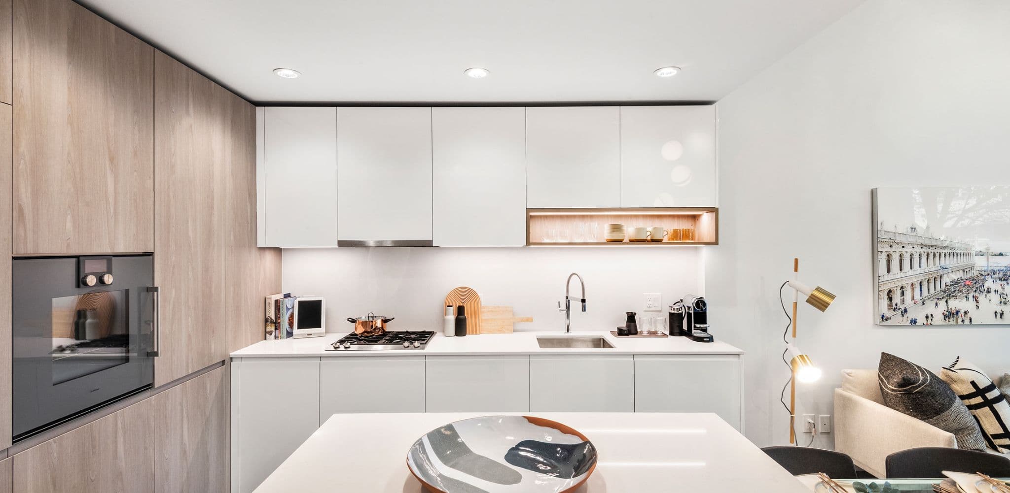 RC at CF Richmond Centre – A modern residential kitchen featuring flat-panel white upper and lower cabinetry paired with light wood grain tall cabinets housing an integrated oven. The space includes bright white quartz countertops, under-cabinet LED lighting, a gas cooktop, and a kitchen island in the foreground with a ceramic decorative bowl.