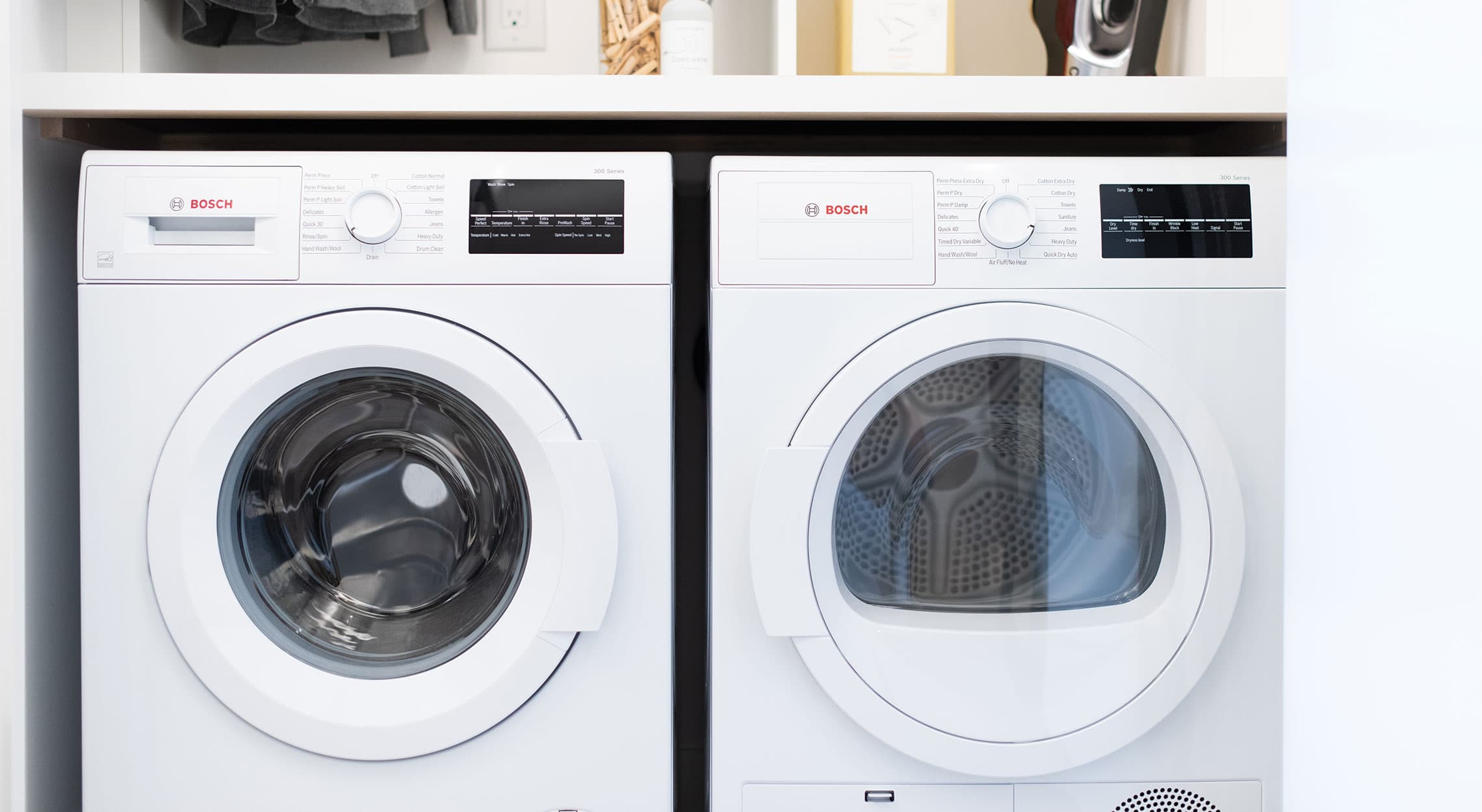 RC at CF Richmond Centre – A close-up view of a modern laundry closet featuring a side-by-side white Bosch front-loading washer and dryer set. The appliances are neatly tucked under a minimalist white countertop with open shelving above holding laundry accessories.