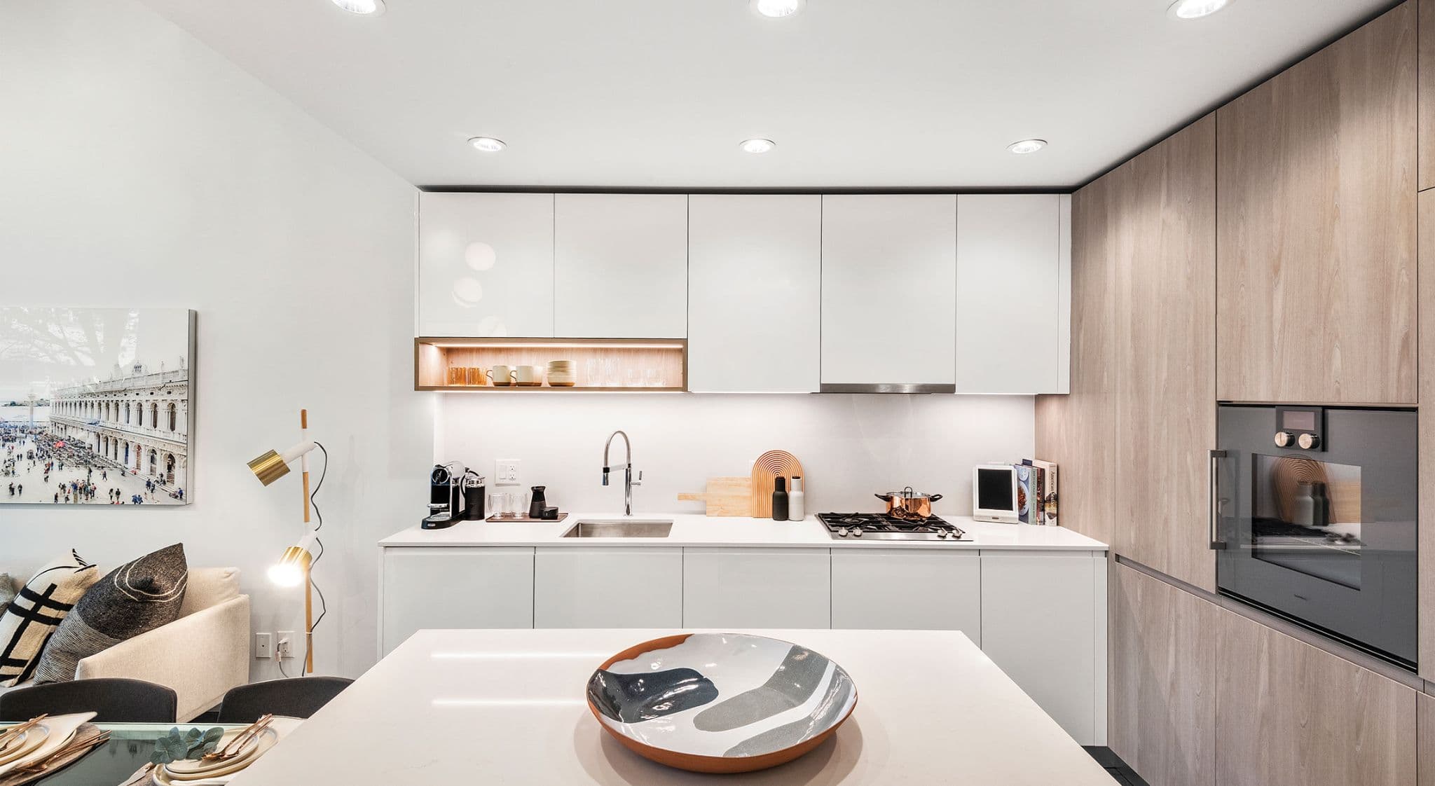 RC at CF Richmond Centre – A modern residential kitchen features clean-lined white flat-panel cabinetry paired with light wood grain tall cabinets and a built-in wall oven. Under-cabinet task lighting illuminates the white countertop, stainless steel sink, gas cooktop, and small counter accessories, while a kitchen island sits in the foreground.