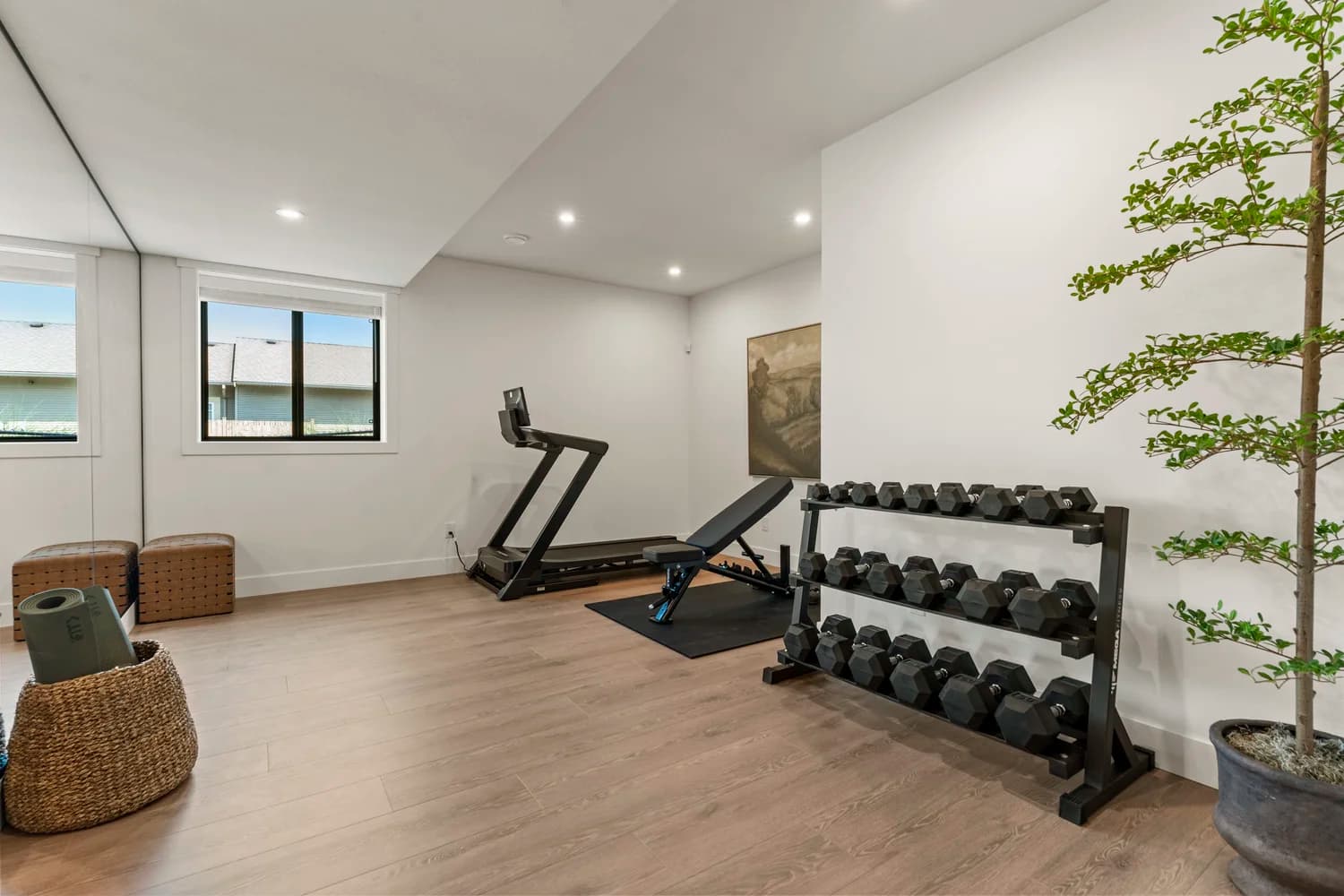Stonewater Ridge – A clean, modern home fitness room features light wood-look flooring, white walls, and a full-length mirrored wall. The space is equipped with a black dumbbell rack, an adjustable workout bench, a modern treadmill, and a tall potted plant that adds a touch of greenery.