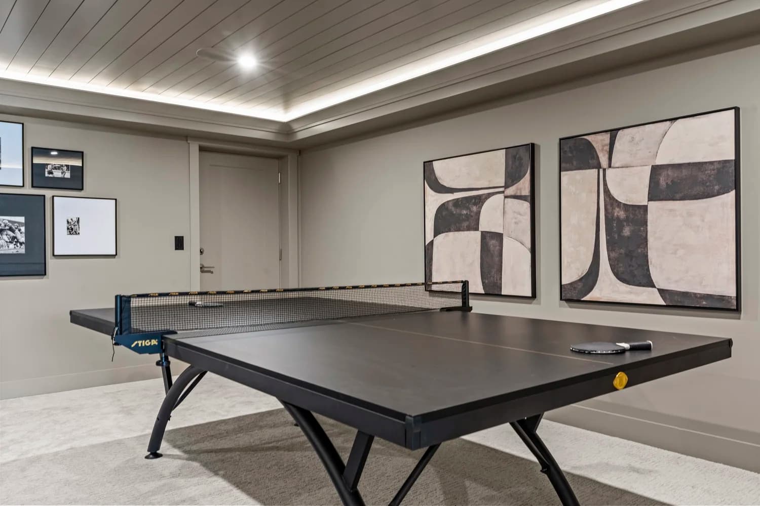 Stonewater Ridge – A modern indoor game room featuring a black Stiga ping-pong table with paddles resting on its surface over carpeted flooring. The space is finished with muted taupe walls decorated with framed black-and-white art prints and modern abstract paintings, accented by a tray ceiling with warm cove lighting.
