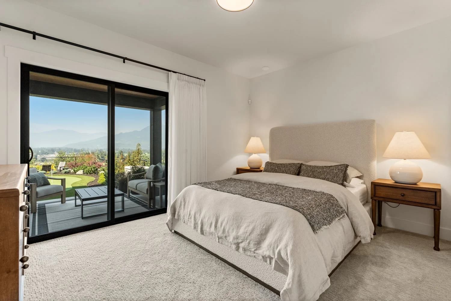 Stonewater Ridge – A bright, neutral-toned bedroom features an upholstered bed flanked by wooden nightstands and soft glowing lamps. Large sliding glass doors open directly to a covered outdoor patio with lounge chairs, overlooking landscaped greenery and distant mountain views.