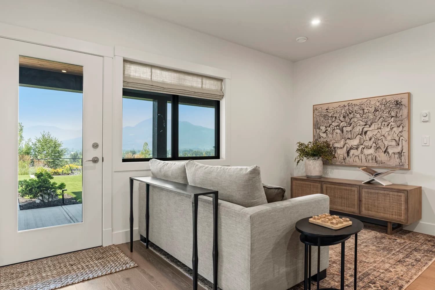 Stonewater Ridge – A bright living room corner features a light grey upholstered sofa backed by a slim black console table, set beside a woven credenza and framed artwork. Glass patio doors and a black-framed window look out onto a manicured green lawn and hazy mountain views in the distance.