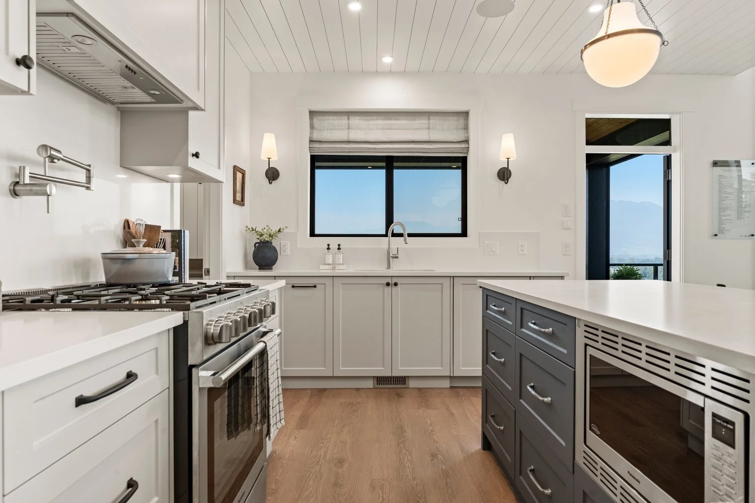 Stonewater Ridge – A bright, modern farmhouse-style kitchen featuring white shaker cabinetry, light wood floors, and a contrasting charcoal-colored island with a built-in microwave. Stainless steel appliances include a gas range with a pot filler, set beneath a shiplap ceiling with wall sconces framing a window that overlooks mountain views.