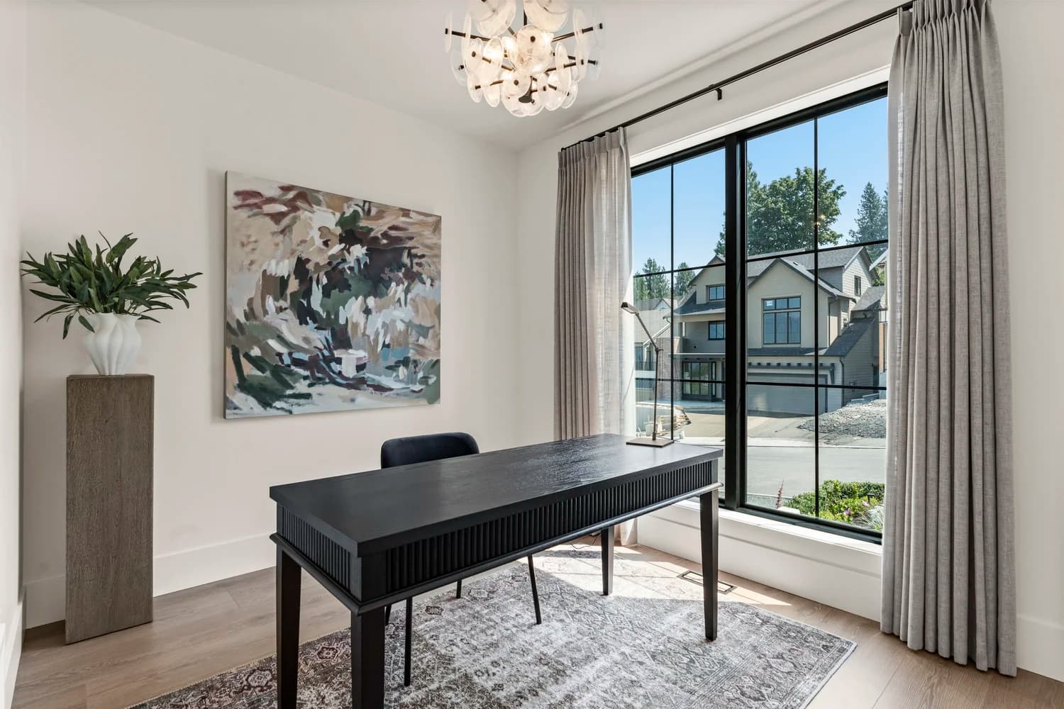 Stonewater Ridge – A bright home office features a dark fluted-wood desk, an area rug over light hardwood floors, and a large multi-pane window framing a suburban streetscape. White walls are accented by an abstract canvas, a sculptural plant on a pedestal, and an organic glass chandelier hanging from the ceiling.