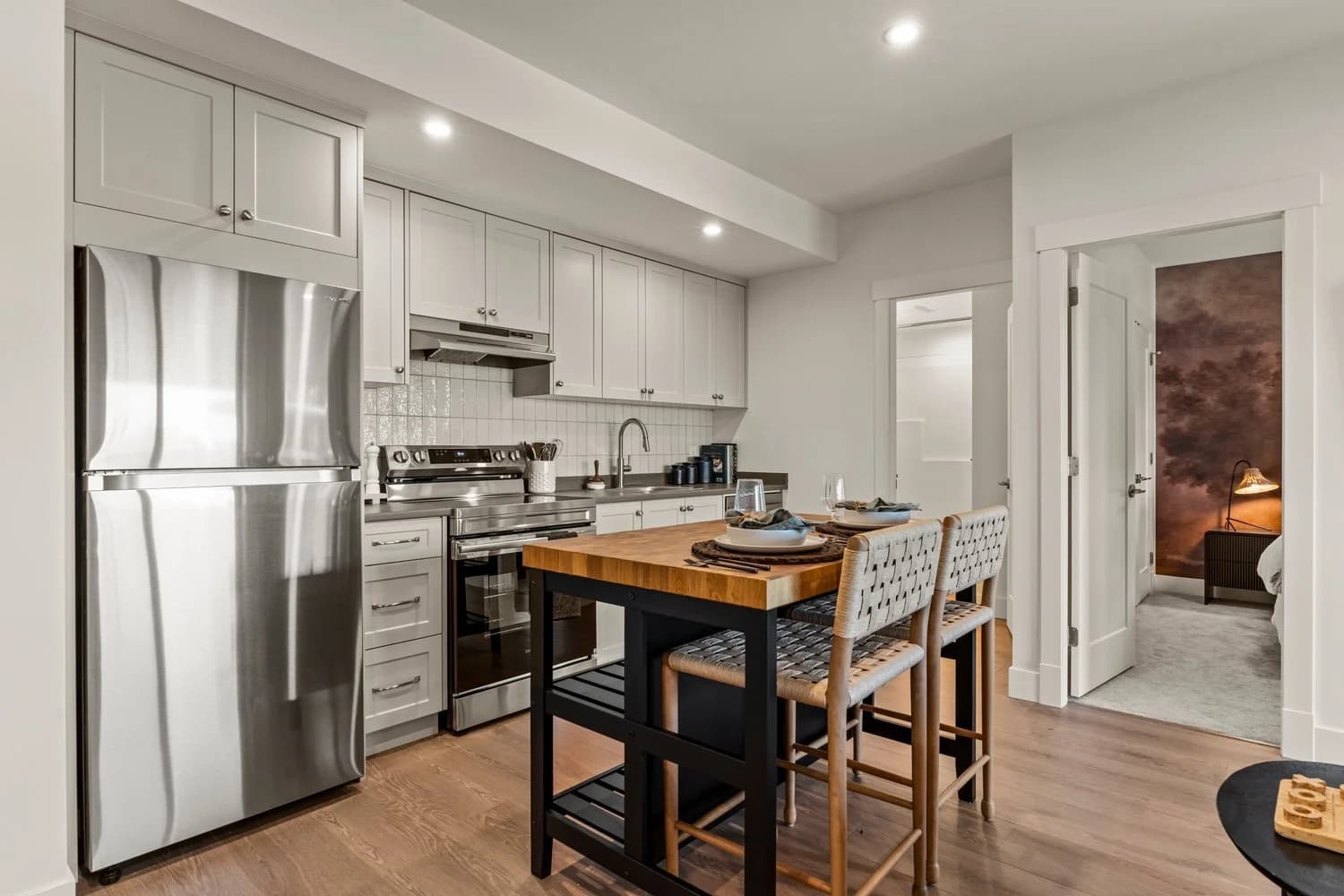 Stonewater Ridge – An open-concept modern kitchen features light grey shaker cabinets, white subway tile backsplash, and stainless steel appliances alongside a butcher-block island with woven barstools. In the background, doorways lead to an adjoining bathroom and a bedroom accented with a dark pictorial feature wall.