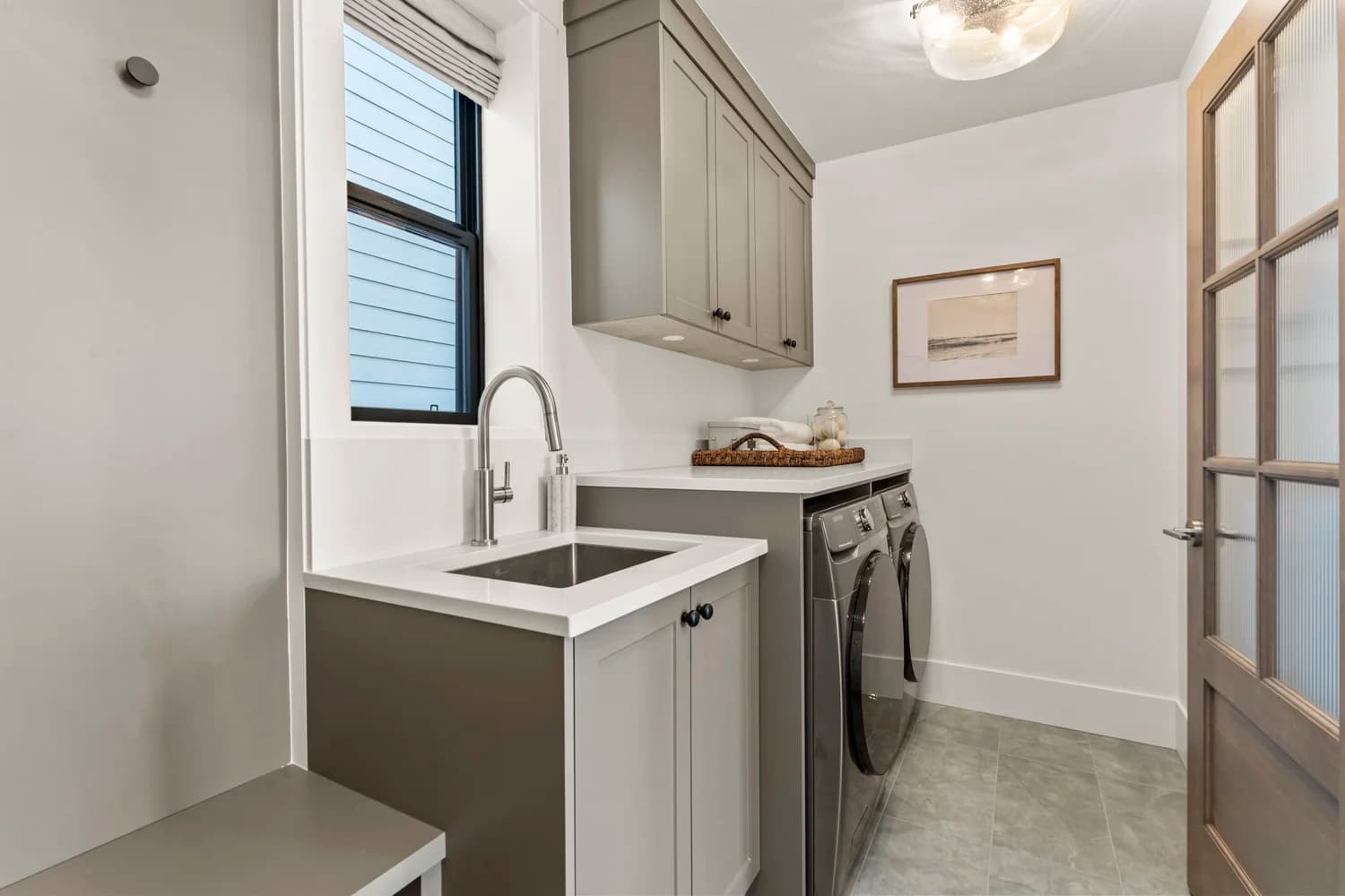 Stonewater Ridge – A clean, modern laundry room features front-loading metallic washer and dryer units beneath a white countertop, alongside a stainless steel utility sink with a gooseneck faucet. Soft grey-taupe cabinetry hangs overhead and below the counters against white walls, accented by a narrow window, grey tile flooring, and a frosted-glass wood door.