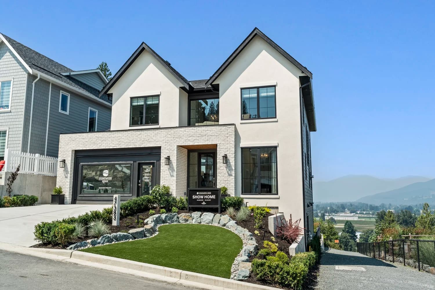 Stonewater Ridge – A modern two-story show home features a bright off-white stucco facade with dual gabled rooflines, black-trimmed windows, and a lower level faced with light-colored brick veneer. Set along a sloped hillside under a clear blue sky, the front yard includes manicured turf framed by a rock retaining wall and offers expansive valley and mountain views in the background.