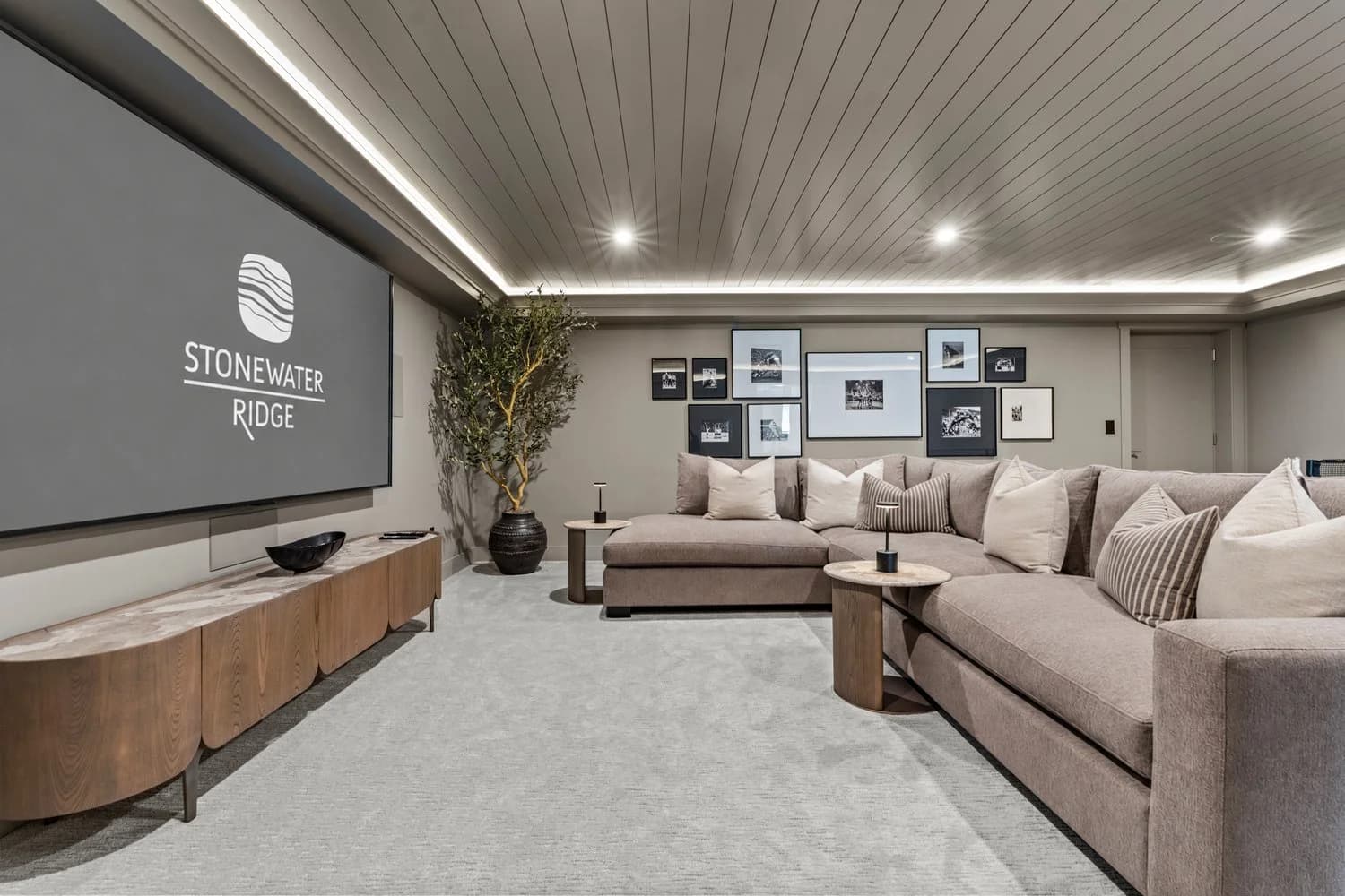Stonewater Ridge – A cozy, modern media room features a large projection screen flanked by a curved wood credenza and an expansive plush sectional sofa accented with throw pillows. Subtle cove lighting illuminates the paneled ceiling, complemented by neutral carpeting, a framed gallery wall, and a potted tree in the corner.